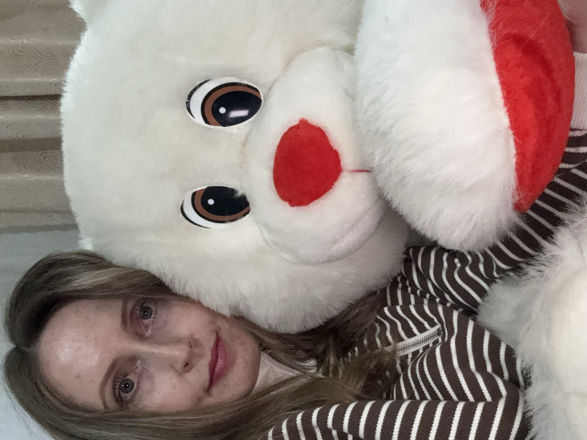 A woman poses for a close-up selfie alongside a large, white plush toy. The image is oriented diagonally, with both subjects filling the frame.
The woman, with fair skin, light-colored eyes, and long, light brown hair, is positioned on the left side of the frame, looking directly at the camera with a subtle, serene expression. She wears a casual garment, likely a jacket or hoodie, featuring horizontal brown and white stripes and a visible silver zipper pull on the collar area. A hint of pink lipstick is visible on her lips.
To her right, the large plush toy, resembling a bear or similar furry creature, dominates the top and right portions of the image. It has big, round brown eyes with white highlights, and a prominent, oval-shaped red nose. A red Santa hat is partially visible on the upper right side of its head, suggesting a festive or playful context. The plush toy has soft, white fur.
The background appears to be an indoor setting, indistinct but suggesting a comfortable environment, possibly a bed or sofa. The lighting is soft and even, indicating an indoor setting without strong natural light. No specific time of day or weather is discernible. There is no visible text in the image. The scene conveys a relaxed and cozy atmosphere. This media file was captured in Chaikovsky, Russia.