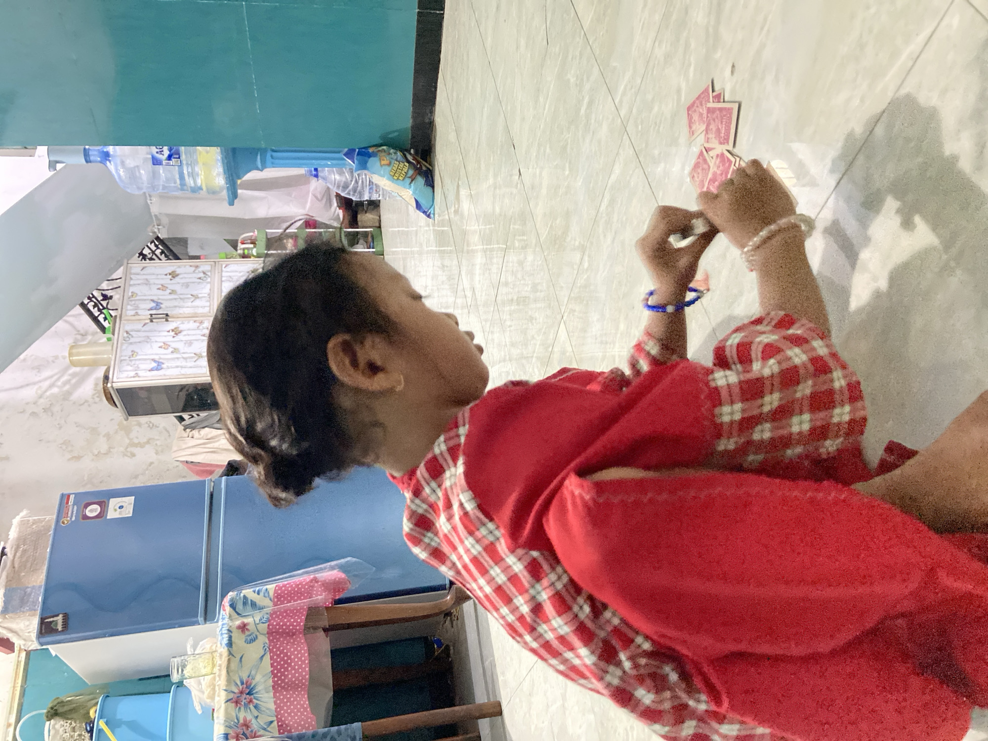 A young child is depicted playing with small, pink cards on a light-colored tiled floor inside a home in Gelam, Indonesia. The child is seated on the floor, facing towards the left side of the frame, with their profile visible. They are dressed in a red and white plaid long-sleeved shirt layered under a plain red garment, possibly a dress or a smock. Both wrists are adorned with beaded bracelets, one white and one blue.
The central activity shows the child actively engaged with the cards. Their left hand holds one card, while their right hand is positioned near a small pile of scattered cards on the floor, suggesting a game or sorting activity. The floor features a faint diamond-grid pattern.
The background provides glimpses of a domestic environment. To the left, a blue two-door refrigerator stands against a wall. Above and behind it, a blue water dispenser holds a large plastic bottle with "AQUA" text visible on its label. A blue package, possibly containing food items, rests on the floor in the mid-ground, showing partially legible Indonesian text including "KEMAS" and "KEPALA." A light-colored cabinet with a decorative, possibly floral or bird, pattern is also visible. The walls are painted in hues of light blue and white. The indoor lighting suggests it is daytime. The child appears focused and absorbed in their play.