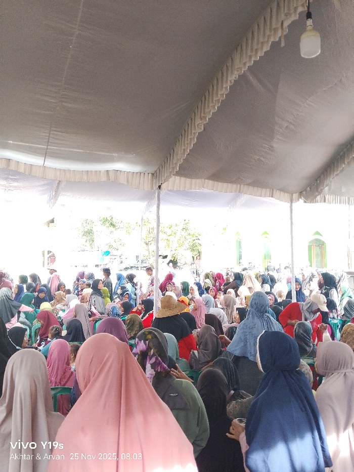 A large crowd of people, predominantly women wearing headscarves of various colors, are seated under a white tent. The scene is brightly lit, with natural light entering from beyond the tent's edges. A single light bulb hangs from the tent's ceiling. The event appears to be taking place outdoors, possibly adjacent to a building with a green window frame. The photo includes a watermark with the device model and date.