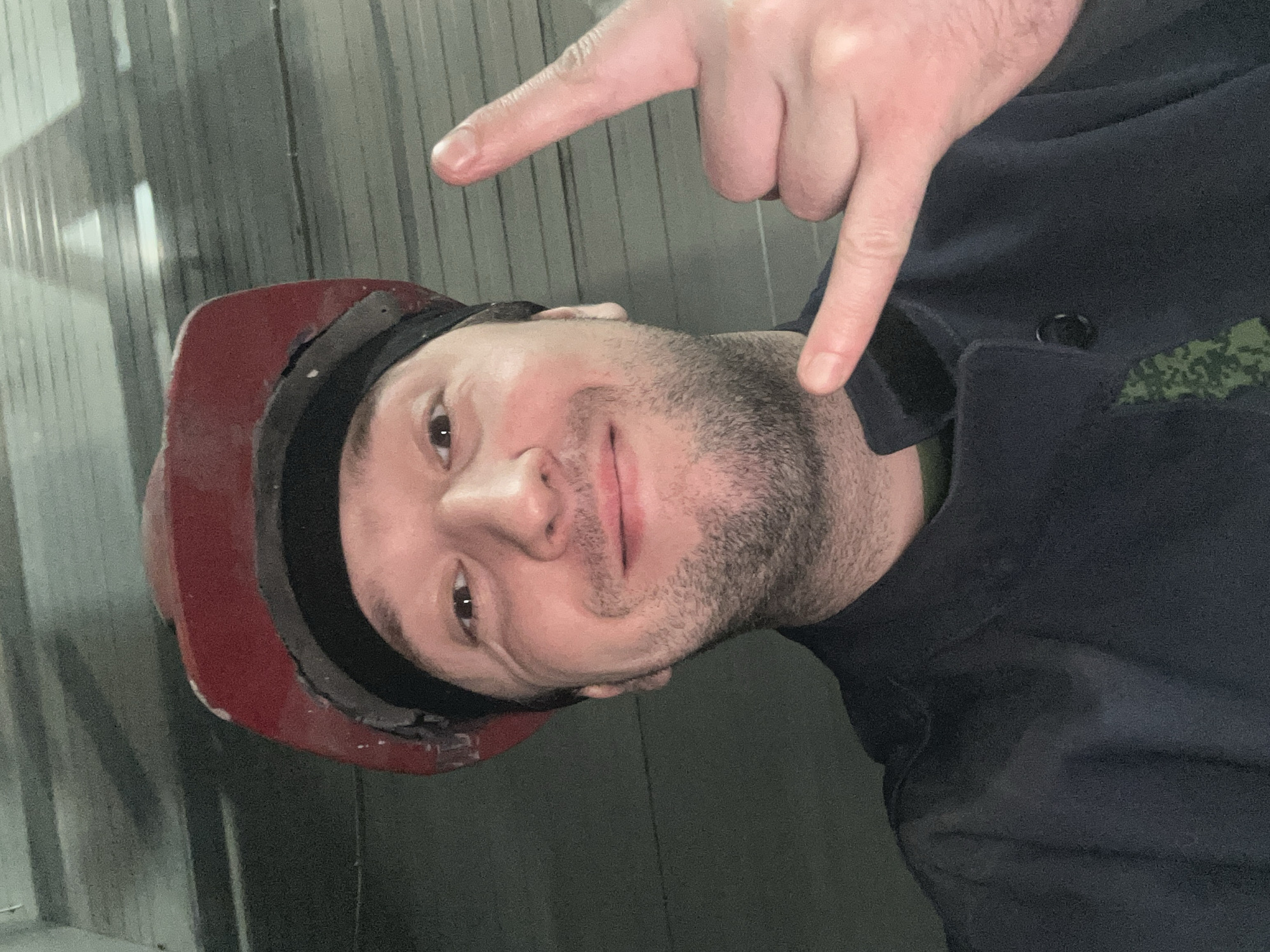 A man is captured in a close-up, slightly rotated portrait, likely a selfie. He has a light complexion, dark short hair, and a beard or heavy stubble, with a faint, confident smile. He is wearing a red hard hat, which shows signs of wear and scuffs, and a dark-colored uniform jacket. A green, pixelated camouflage pattern patch is visible on his right shoulder or upper arm area. A hand, likely his own, is positioned above his head, with two fingers pointing downwards towards his body. The background consists of vertical, corrugated metal or paneling, suggesting an indoor industrial, workshop, or service environment. The lighting is somewhat dim. No text is visible. The provided context indicates the location is Noviy, Russia.
