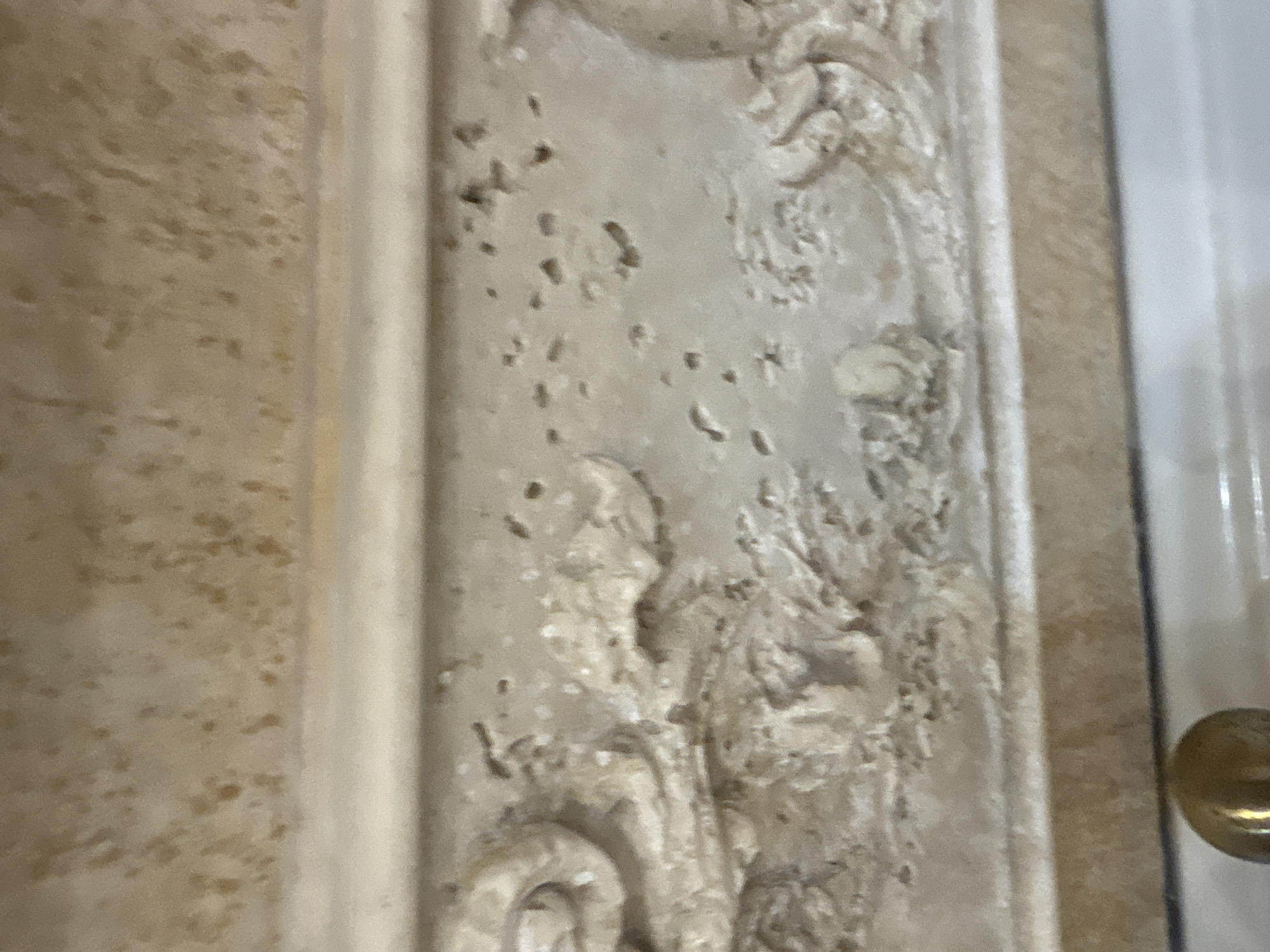 This is a close-up, angled, eye-level photograph of a light-colored, textured surface with carved detailing. The surface appears to be stone or a similar material, possibly a decorative architectural element or furniture. It has a porous texture with small pits and irregular darker speckles throughout. A raised band runs horizontally across the mid-section of the image, featuring ornate, swirling carved designs on the left side. Below this band is a flatter, lighter surface. The top portion of the image shows a more textured, uneven surface with a mottled appearance. A small, metallic, spherical object, possibly a knob or finial, is visible in the bottom left corner, reflecting light. The overall lighting suggests an indoor environment. The image does not contain any people or provide any context for its location within Georgiyevsk, Russia.