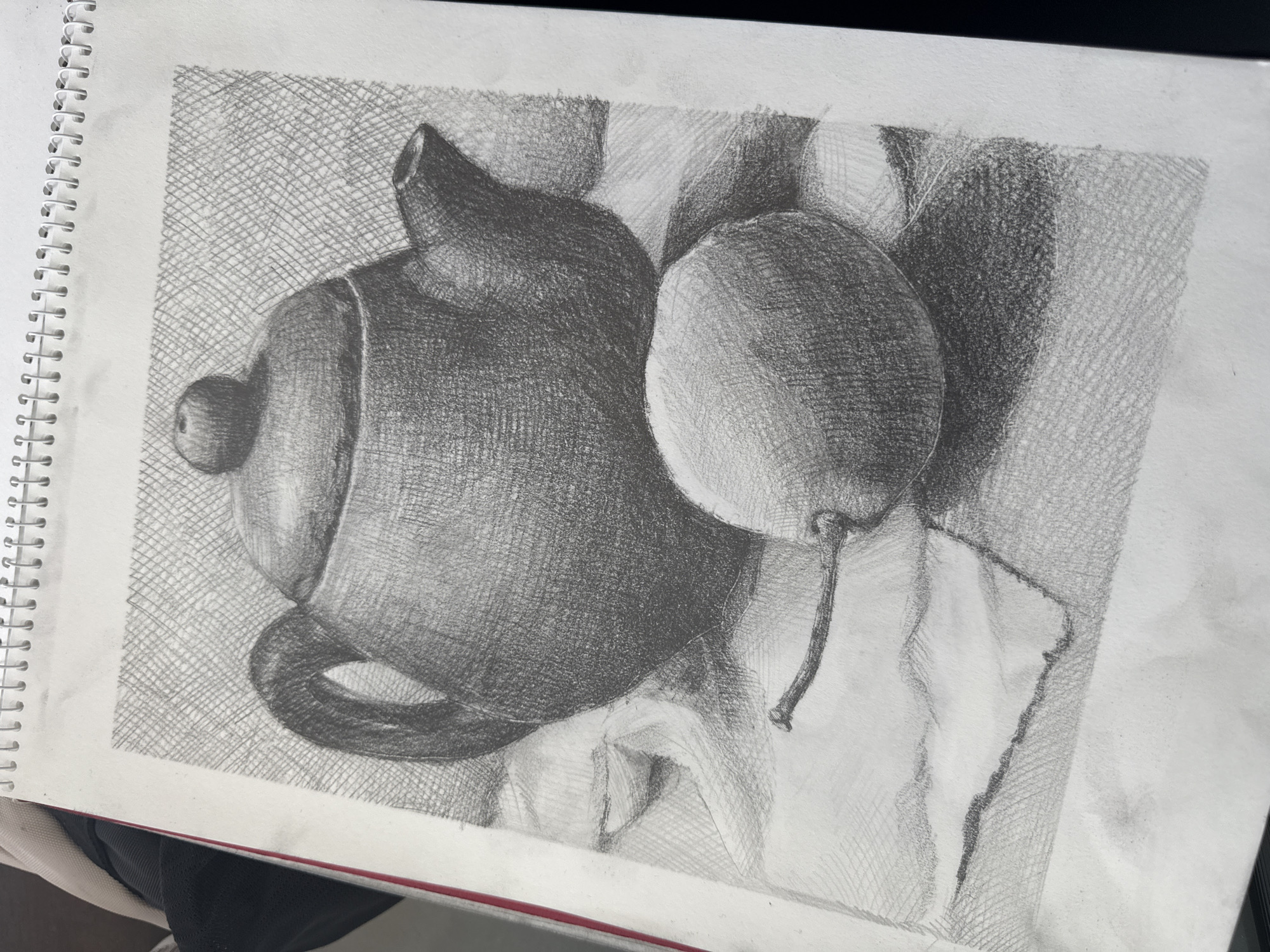 A pencil drawing on a spiral-bound sketchbook depicts a still life. The objects include a teapot and a pear. The background is a cross-hatched pattern. The shading on the teapot indicates a dark, matte finish, while the pear has a lighter, reflective surface. The placement is inside or in a personal place. The description does not specify it is located in the city of Sofia, Bulgaria.