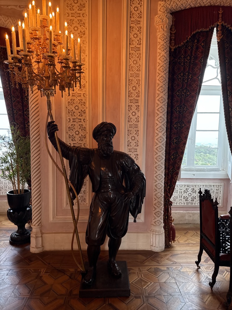 The image depicts an interior scene, likely within Pena Palace in Sintra, Portugal, suggested by the ornate decor and architectural details. A bronze statue of a man wearing what appears to be Ottoman-style clothing stands prominently in the foreground. The statue holds a long, curved staff or pole, from which a multi-tiered chandelier is suspended, lit with numerous candles.
The room features patterned wood flooring, pinkish-peach walls with detailed trim, and tall, narrow windows draped with heavy, maroon patterned curtains. An ornamental potted plant stands in the left background, and a portion of an ornate chair can be seen on the right. The overall environment suggests an opulent, historical setting.