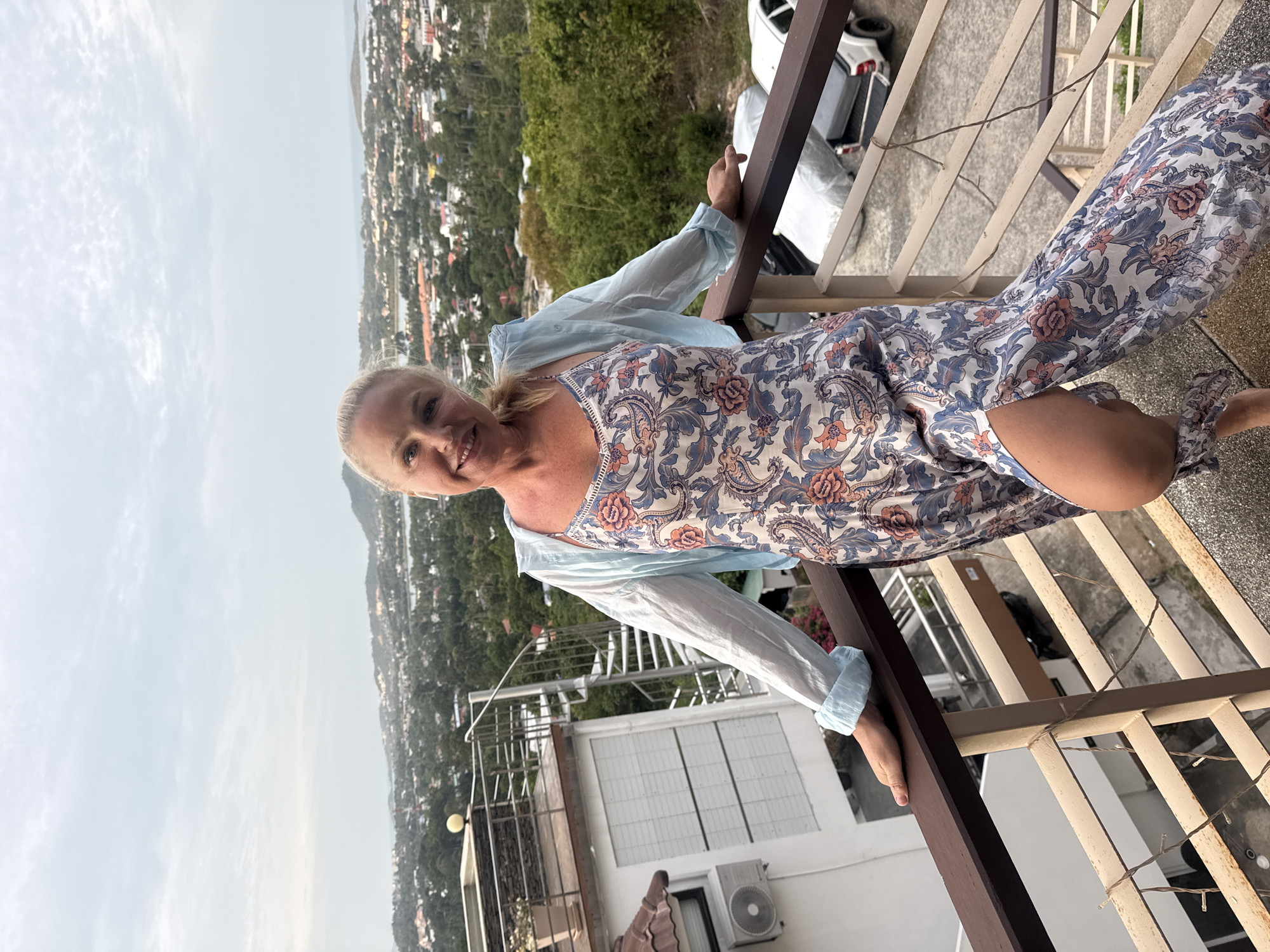 A blonde woman stands on a balcony overlooking a city and ocean vista. She is wearing a floral-patterned dress and a light blue open shirt. The balcony has a brown wooden railing and beige metal bars. In the background, the city is dense with buildings and greenery, leading to a body of water. The sky is overcast. The location appears to be Ko Samui District, Thailand.