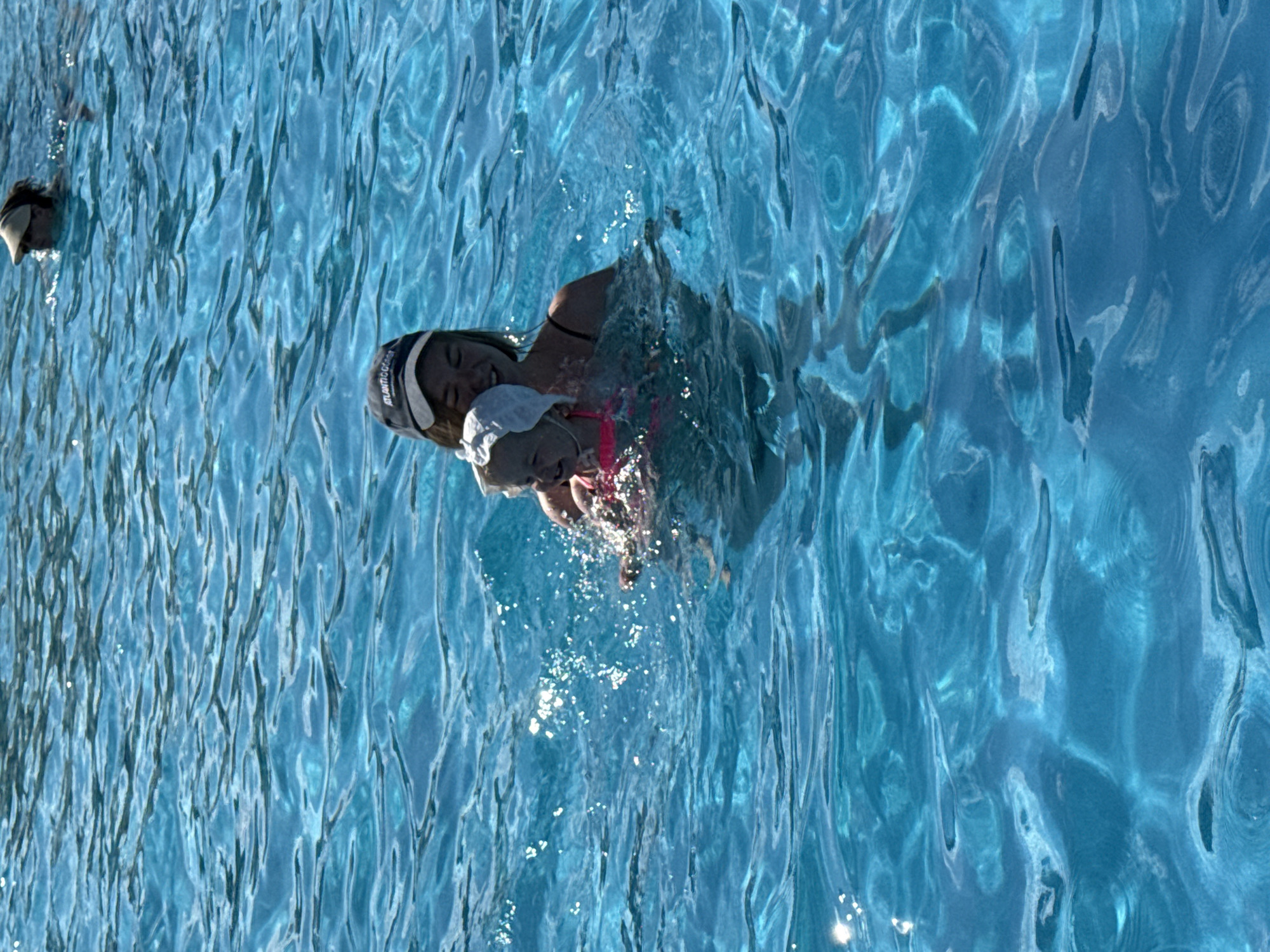 In the city of Koloksha, Russia, a young woman wearing a blue baseball cap and a black swimsuit is in a swimming pool, holding a toddler. The toddler is wearing a white cap and a pink swimsuit. Both are partially submerged in the clear blue water, with ripples and sunlight reflecting on the surface. Another person, wearing a sun visor, is visible swimming in the background of the pool. The scene depicts a recreational activity in a public swimming facility.