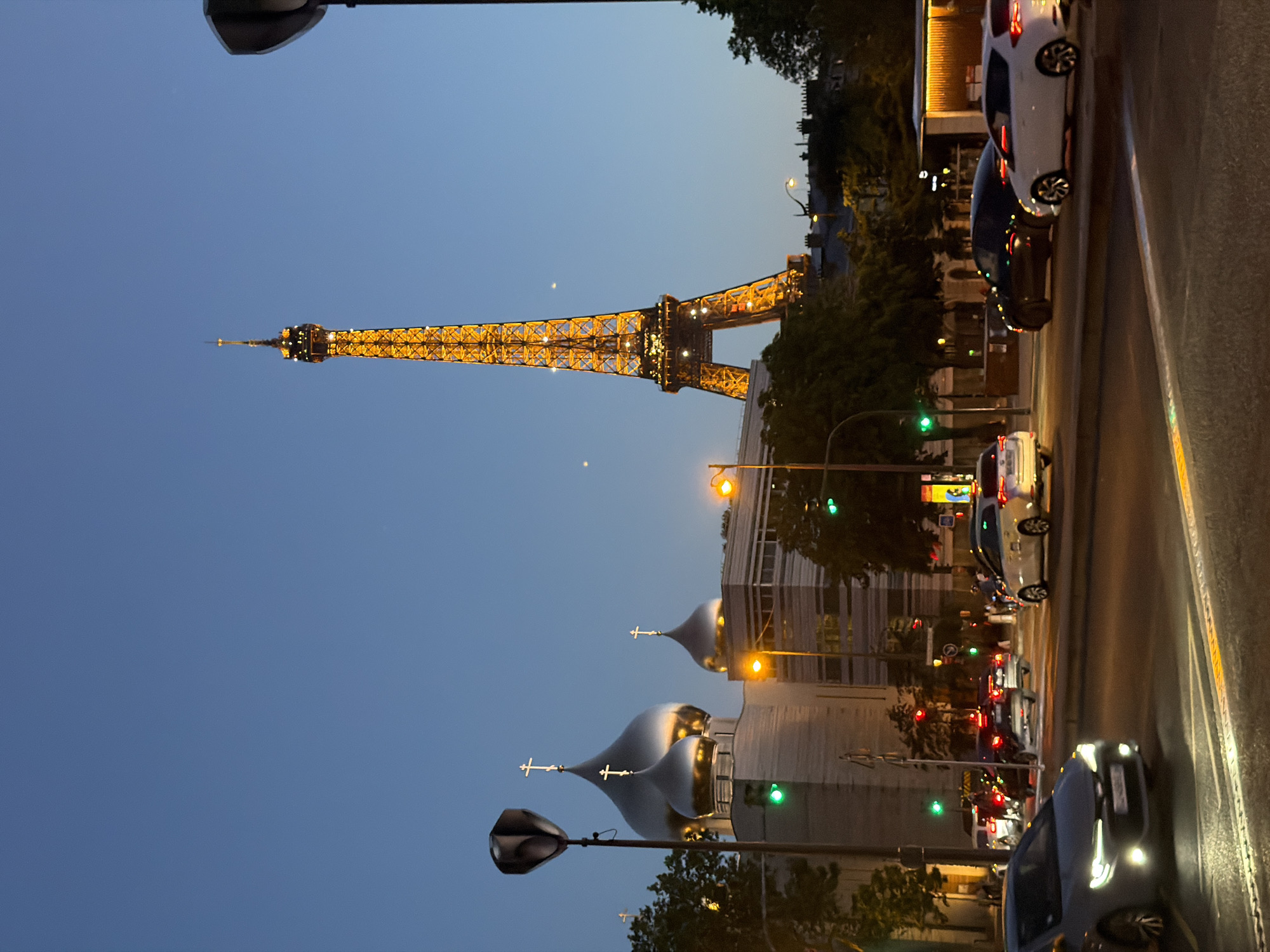 This image captures a street scene in Paris, France, at dusk or night. The Eiffel Tower, illuminated with yellow lights, dominates the background sky. In the foreground, the silver domes of the Russian Orthodox Cathedral are visible, each topped with a cross. Streetlights illuminate the scene, and traffic is present, with cars parked along the street. The architecture of the surrounding buildings reflects a mix of modern and traditional Parisian styles. The sky is a gradient of dark blue, indicating the transition from day to night. The composition is slightly tilted, suggesting the photo was taken spontaneously.