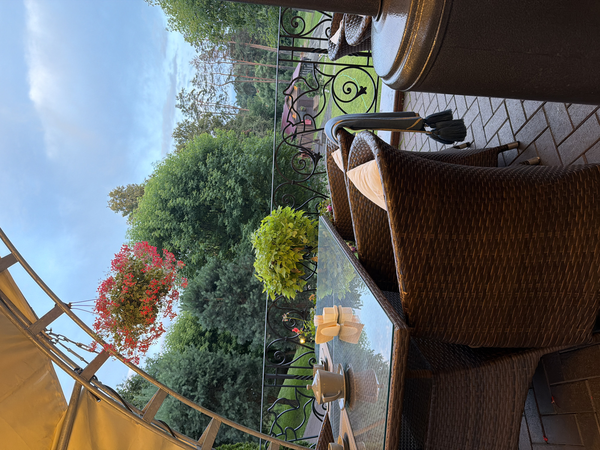 The image depicts an outdoor seating area at a restaurant or cafe, likely located in Izmailovo, Russia, given the presence of a dacha-style building in the background and lush greenery characteristic of the region. The scene is set on a patio paved with rectangular stone tiles. In the foreground, a glass-topped table is set with a teapot, cups, and a napkin holder, suggesting a tea or coffee service. Wicker chairs with cushions surround the table. Above the seating area, a fabric awning is suspended by a metal frame, and a hanging basket filled with red flowers dangles from it. Beyond the patio, a decorative wrought-iron railing overlooks a grassy area with trees and a rustic wooden building with a red roof. The sky is overcast, with a soft, diffused light. No people are visible in the frame.