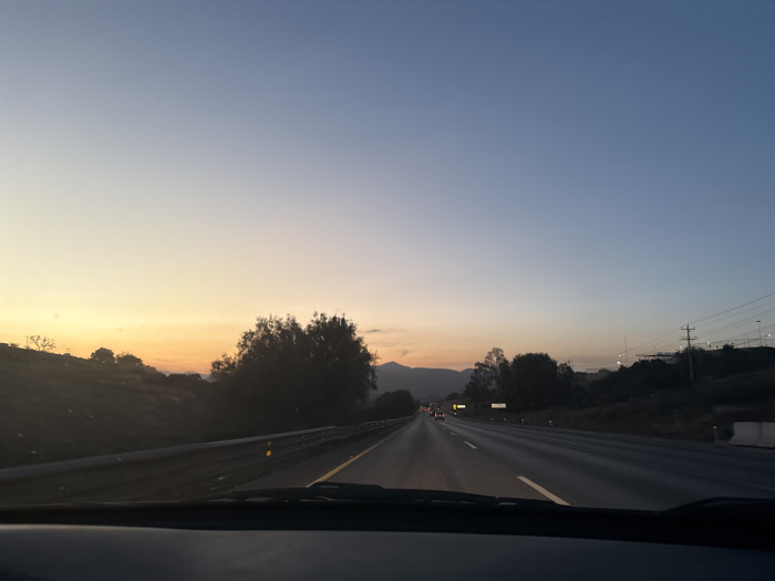 A view from a car driving on a highway at dusk. The sky transitions from yellow to blue. The road stretches ahead, showing a few cars in the distance. Mountains are faintly visible on the horizon. Guardrails line the road's edge, and trees are present on the adjacent hillsides. Power lines are visible on the right. The car's dashboard is in the foreground.