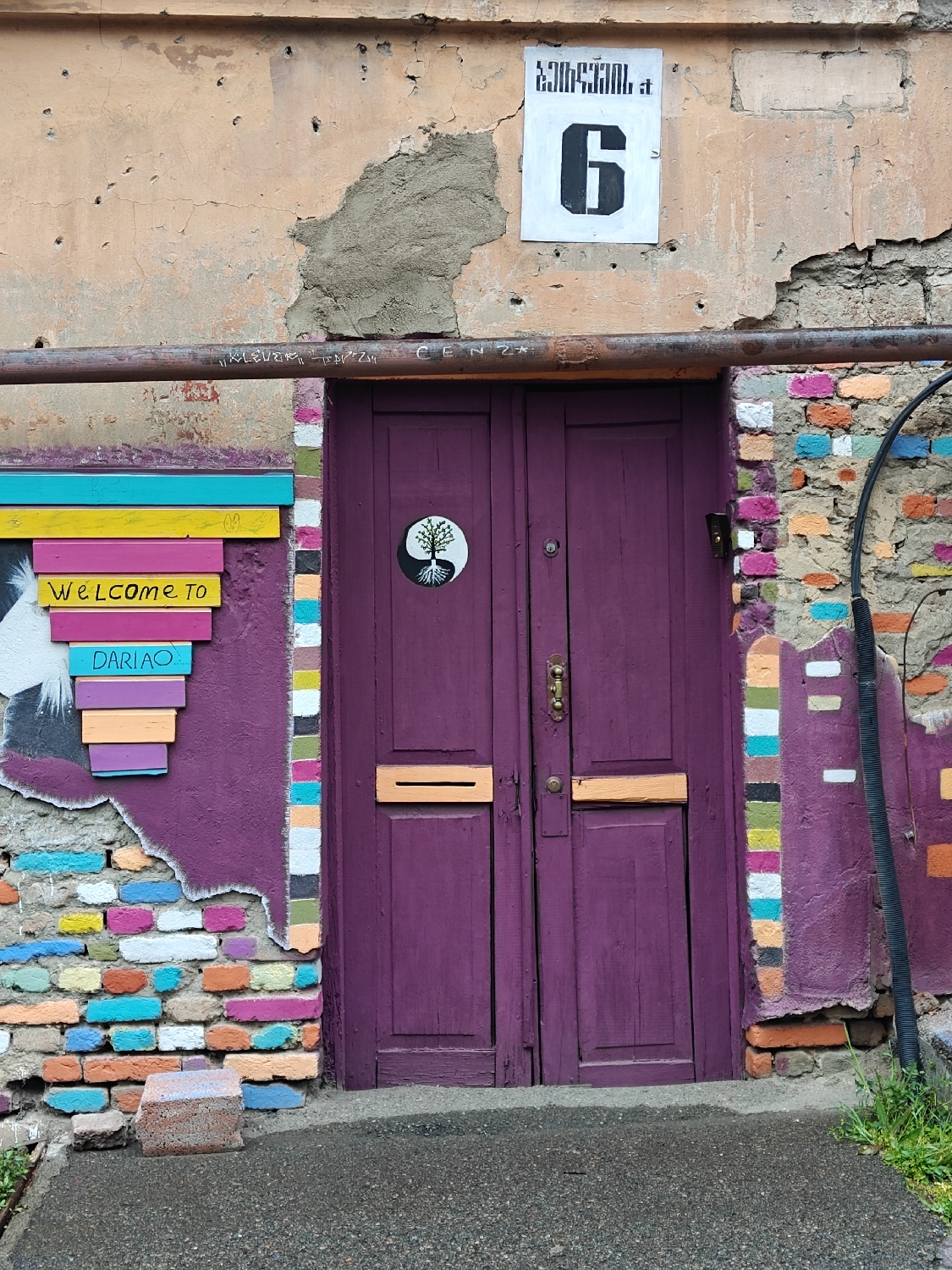 This image captures a vibrant and eclectic street scene, characteristic of Tbilisi, Georgia. The central focus is a deeply purple, double wooden door, adorned with brass-colored hardware and two horizontal orange-painted mail slots. A circular black and white symbol resembling a yin-yang, with a tree design, is fixed to the left door panel.
Above the door, a white rectangular sign displays the bold black number "6," with Georgian script (ქართული) visible above it. The wall surrounding the door is a canvas of textures and colors. The upper section features a light orange, peeling facade with numerous indentations and a rough concrete repair patch. Below this, the wall transitions into a striking purple, which partially reveals an underlying brick wall where individual bricks are painted in a kaleidoscope of bright colors, including blue, pink, orange, yellow, and white. This colorful brickwork extends along both sides of the door.
To the left of the door, a series of horizontally stacked, brightly painted wooden slats spell out "Welcome to DARIAO" in white stenciled letters. A rusted metal pipe runs horizontally across the upper wall, featuring graffiti that includes "KLEVER" and "SENZA." A black pipe descends vertically along the right side of the door. The ground in front is concrete, with a few loose, painted bricks at the base of the wall and a small patch of green grass on the far right. The scene, captured in daytime under soft lighting, depicts a static architectural detail that blends urban decay with artistic expression, creating a welcoming and visually stimulating atmosphere.