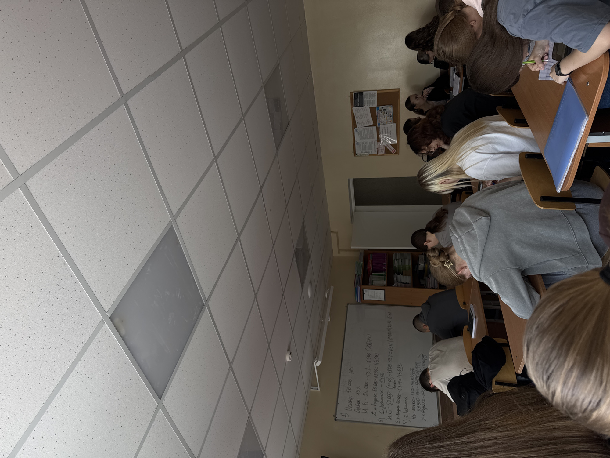 The image depicts a classroom in what appears to be an educational institution, likely a school or university in Perm, Russia. Several students are seated at desks, presumably taking notes or working on an assignment. A whiteboard displays equations and calculations written in Russian. The classroom has a standard drop ceiling with fluorescent lighting. The setting suggests an academic environment where instruction and learning are taking place. A bulletin board is visible on the wall next to the whiteboard.