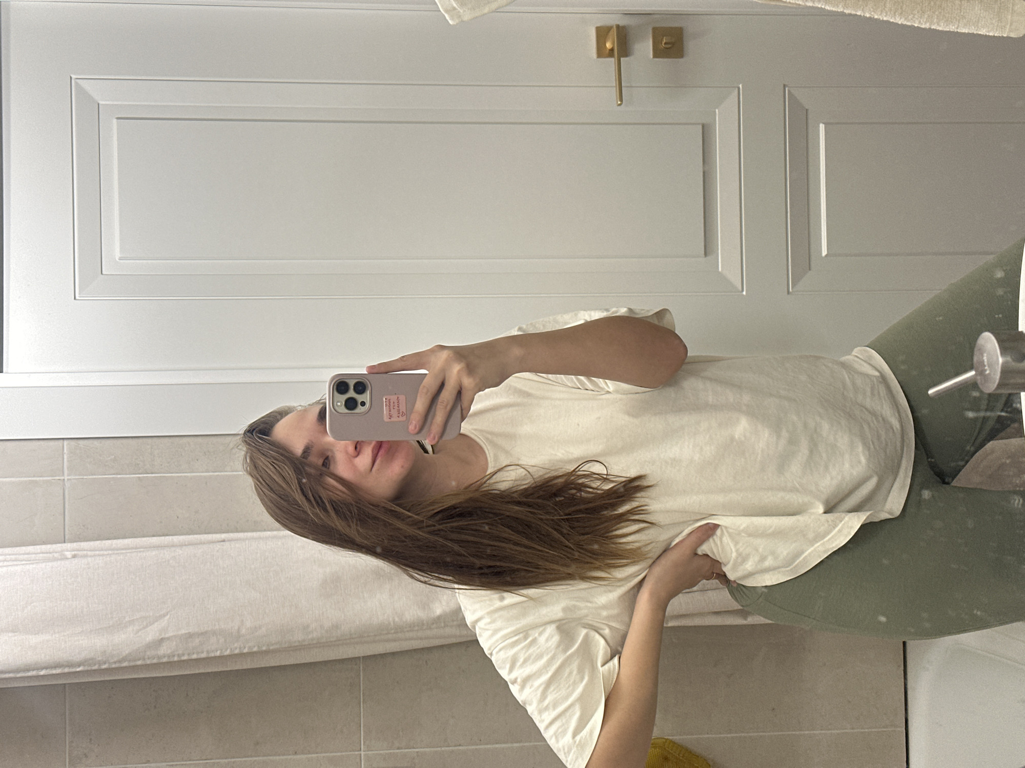 A woman takes a selfie in a bathroom. She is wearing a light-colored t-shirt and green pants. She is holding a pink smartphone. The bathroom has white wall tiles, a white shower curtain, a white door with gold handles, and a sink. The location is not specified.