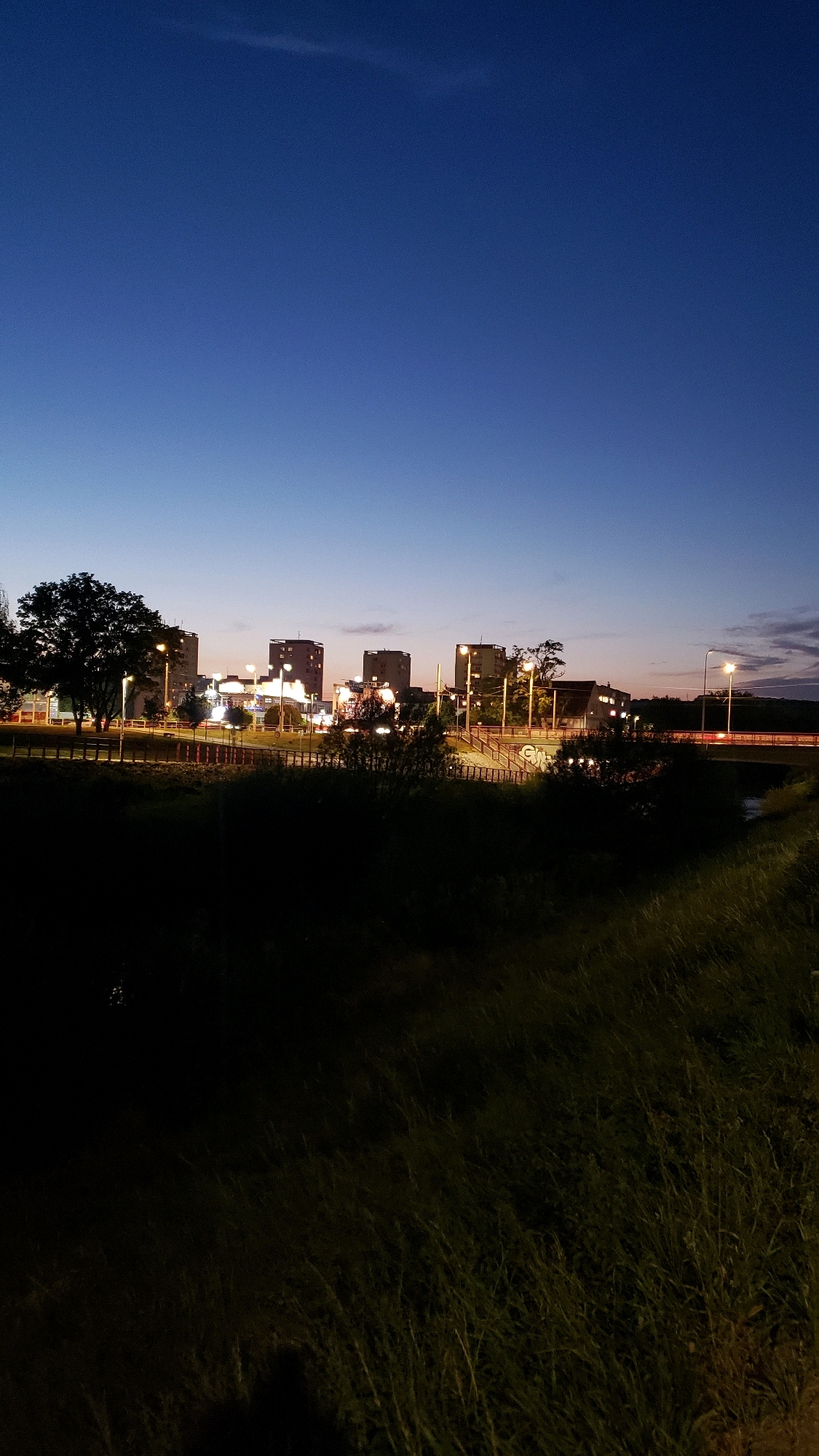 The image depicts a cityscape at dusk, likely in Prešov, Slovakia, featuring a line of buildings and a wide expanse of dark grass in the foreground. The sky transitions from a deep blue at the top to a lighter, warmer hue near the horizon, where the last vestiges of daylight are visible. Several tall buildings, illuminated by artificial lights, dominate the midground. They appear to be residential or office blocks, with windows glowing from within. Streetlights are also visible, casting pools of light on pathways and along a road that runs horizontally across the scene. A bridge or overpass is partially visible in the right midground. The foreground is comprised of a large area of tall, dark grass, suggesting a natural or park-like setting adjacent to the urban development. The overall impression is one of an urban area transitioning from day to night, with the lights of the city beginning to assert themselves against the darkening sky.