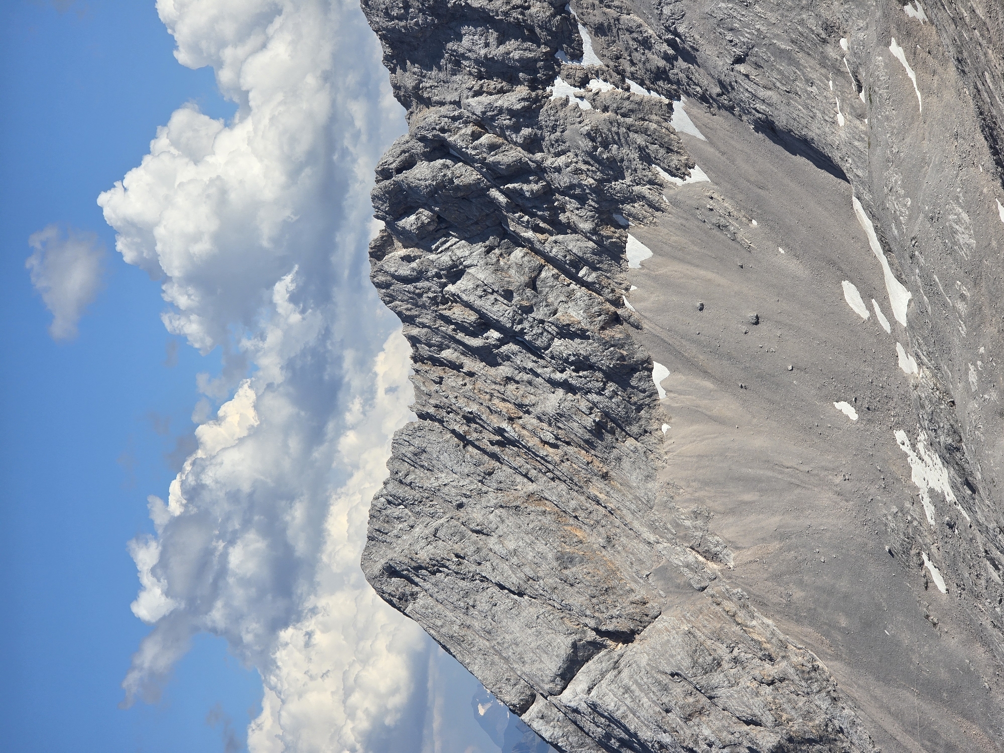 The image depicts a mountain landscape with a rugged, rocky peak under a partly cloudy blue sky. The mountain slopes are covered in scree and patches of snow. The rock formations are characterized by visible layers and fissures, indicating a geological structure. The overall environment is natural and mountainous. This scene is likely from the Zugspitze massif, located in the Wetterstein Mountains on the border between Germany and Austria, and is a prominent natural landmark associated with the city of Garmisch-Partenkirchen, Germany. There are no people or man-made objects visible in the image.