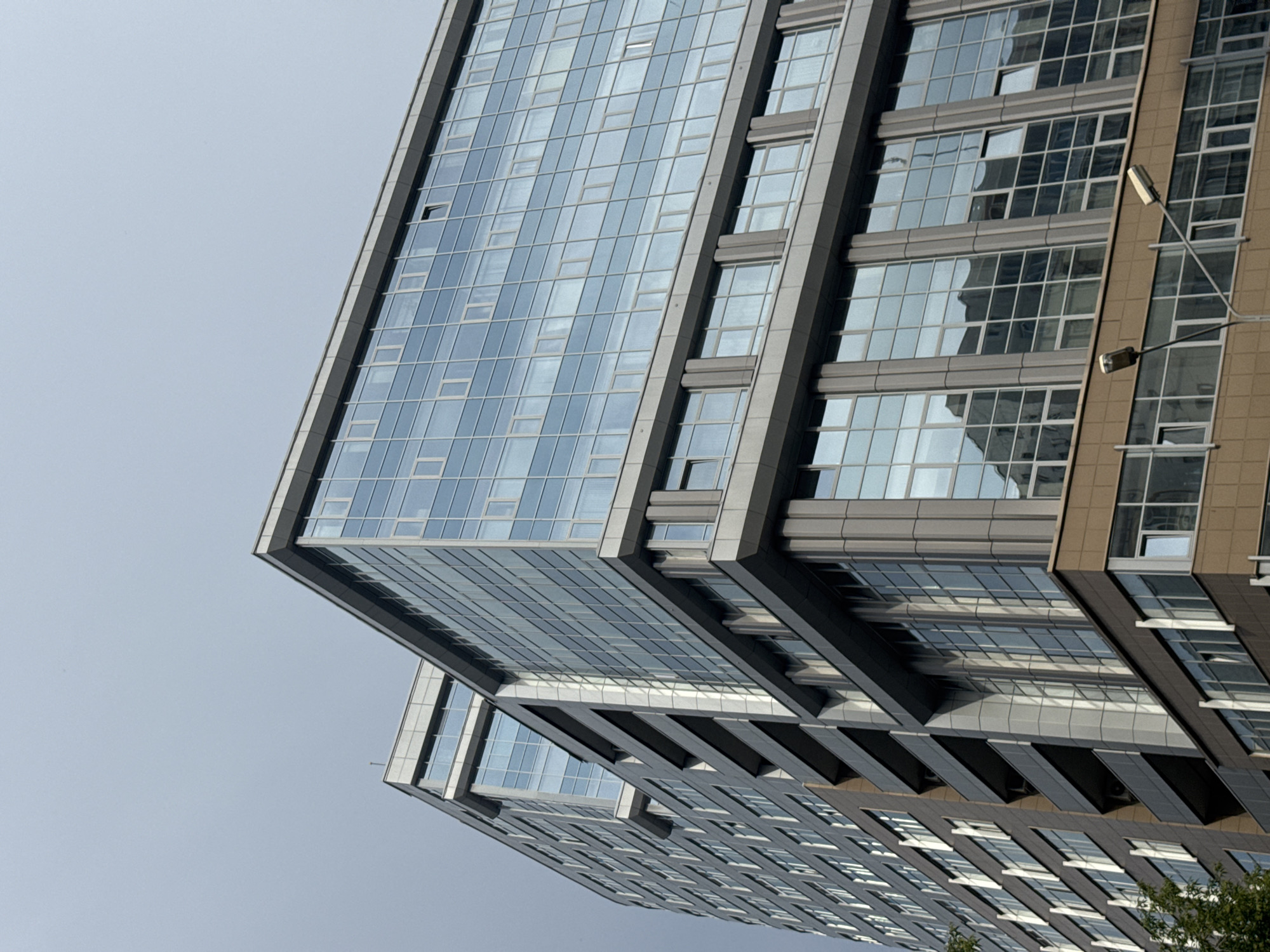 The image shows a modern, multi-story building with a facade primarily composed of glass and grey panels. The building features a grid of windows, many of which reflect the overcast sky. The architecture is geometric and appears to be an office or residential complex. Two streetlights are visible in the foreground, extending from the lower right and center of the image, angled upwards towards the building. The scene is set against a pale, cloudy sky, suggesting an urban environment. The building's design suggests it is located in a contemporary city.