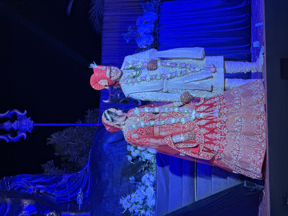 A newlywed Indian couple is seen posing in front of a blue-lit backdrop adorned with flowers and leaves. The bride wears a red lehenga with intricate embroidery, a matching veil, and jewelry. The groom is dressed in a white sherwani and pants, with a red turban. Both wear floral garlands around their necks. The couple stands close, with the bride slightly in front of the groom. A decorative trident is visible on the left side.