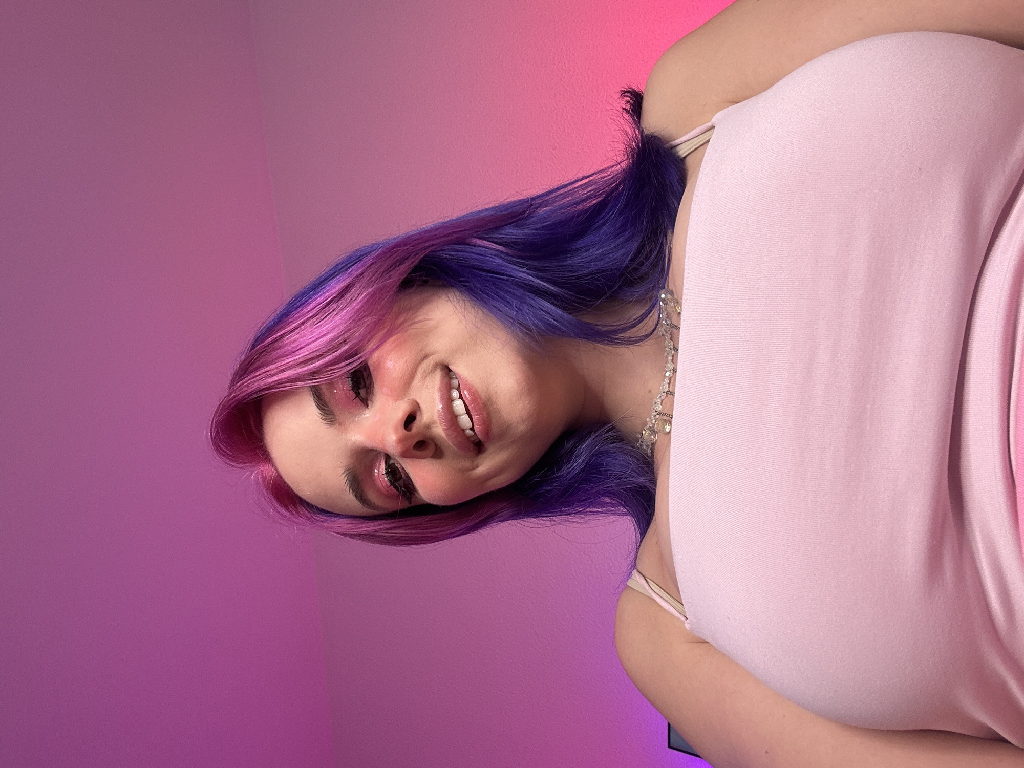 The image shows a young woman with pink and purple dyed hair, wearing a light pink tank top and a silver necklace. She is smiling and looking down at the camera. The background is a gradient of pink and purple light. The scene is a close-up shot, and there is no indication of the location being in Oviedo, Spain.