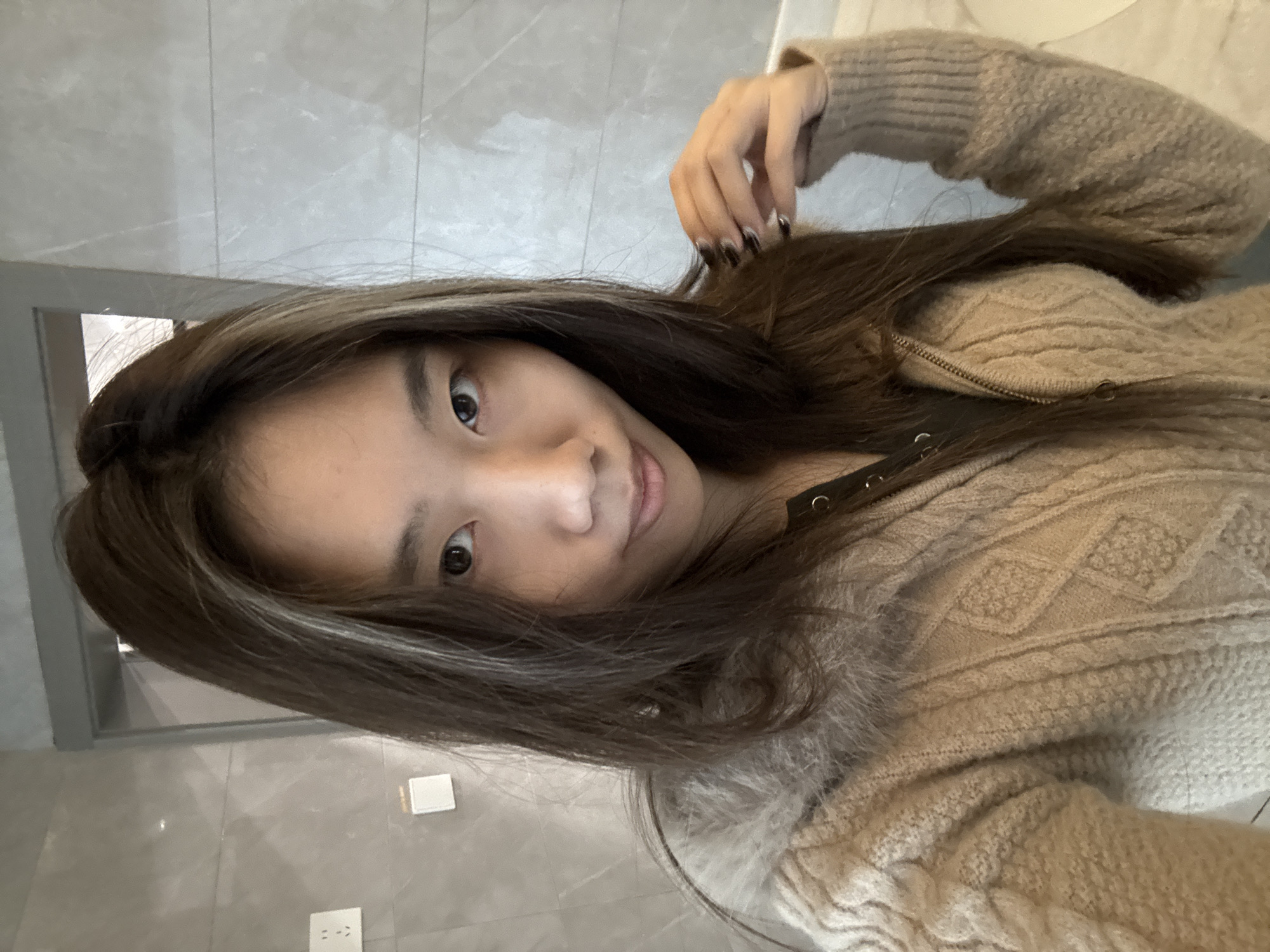 A young Asian woman with long, dark brown hair and lighter highlights poses for a selfie. She is wearing a light brown cable-knit sweater with a fur collar. Her left hand is raised, fingers touching her hair. She is smiling slightly. The background appears to be a tiled wall and doorway, suggesting an interior space such as a bathroom or corridor. The location cannot be determined definitively within the Nan'an District of China based solely on this image.