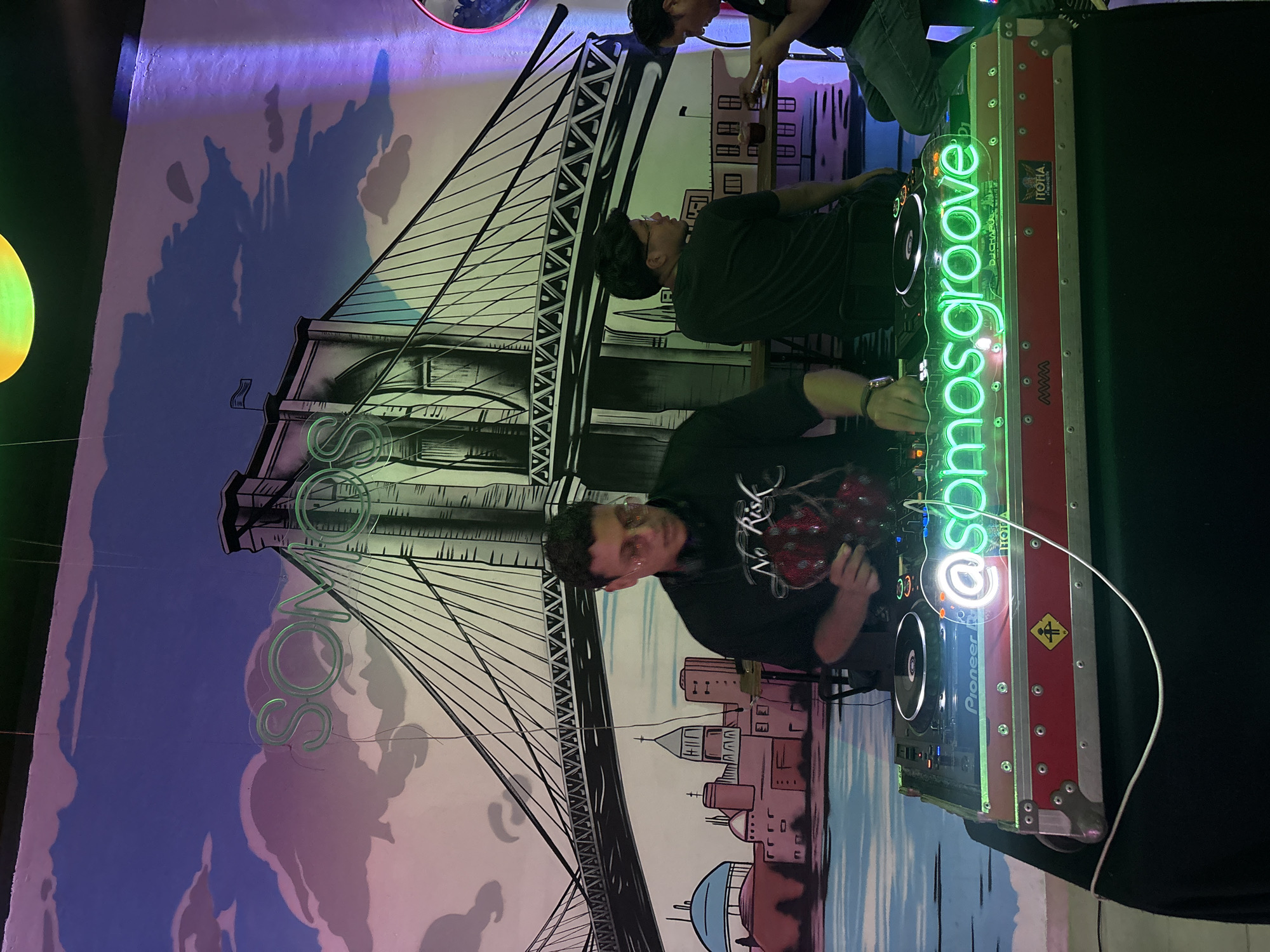 A DJ is performing at a venue with a mural of the Brooklyn Bridge and a generic cityscape behind him. The DJ is a young man wearing a black sweatshirt with a graphic on it. He is operating a Pioneer DJ setup that includes turntables and a mixer. The DJ setup has a neon sign that reads "@somosgroove". Two other people are partially visible in the background. The location is likely an indoor venue in San Bernardino Tlaxcalancingo, Mexico.