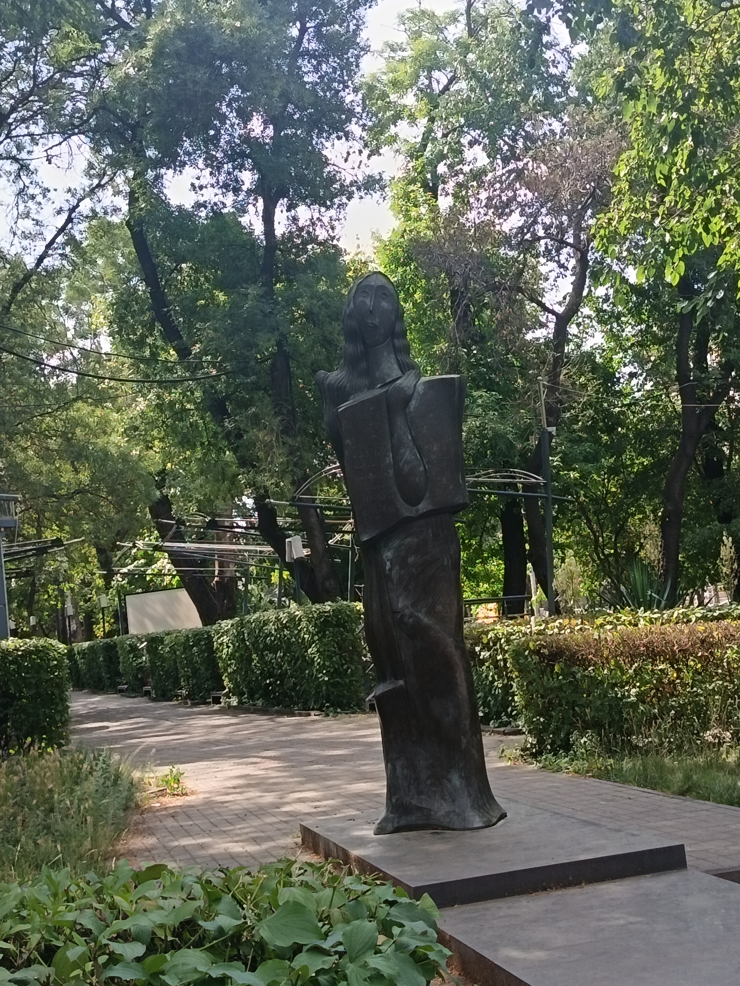 An abstract, bronze sculpture of a figure holding books stands on a concrete pedestal in a park. The sculpture is dark brown and has a smooth, abstract form. The figure's head is tilted slightly to the left, and its face is not detailed. It appears to be wearing a long, flowing robe. The pedestal is rectangular and dark gray.
The sculpture is positioned on a paved walkway made of interlocking bricks. To the left of the walkway, there is a row of neatly trimmed green bushes. Beyond the bushes, there are trees with lush green foliage, suggesting a temperate climate. The background is filled with more trees and vegetation, with sunlight filtering through the leaves, creating dappled shadows on the ground. A white banner or sign is visible in the distance, partially obscured by the trees. To the right of the walkway, there is another row of bushes, some of which appear to be a darker reddish-brown color. The overall environment is serene and verdant, indicative of a public park or garden. The lighting suggests it is daytime, likely in the morning or afternoon, with bright sunshine.