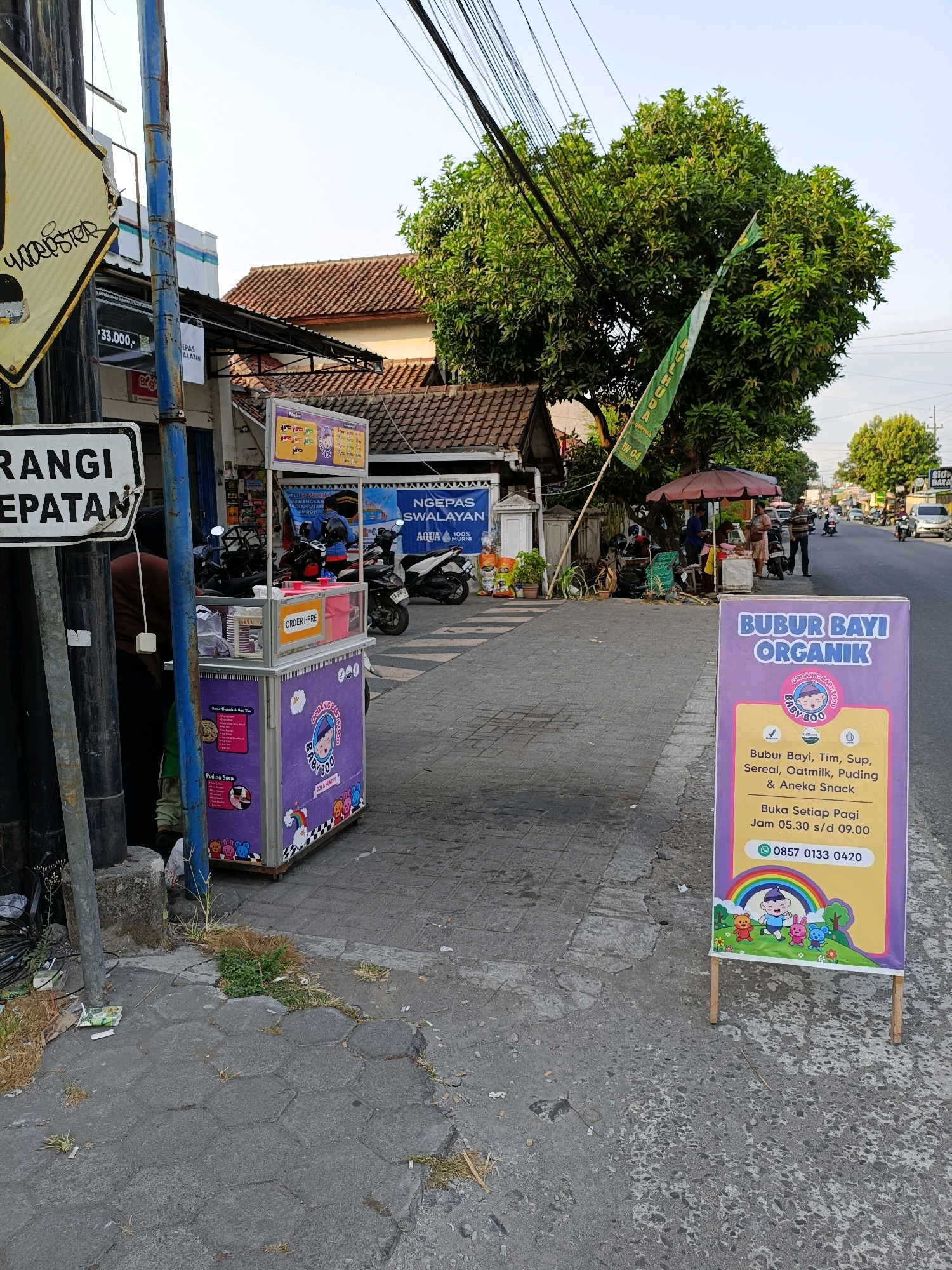The image captures a street scene in Purwomartani, Indonesia, with a focus on a street food cart selling organic baby porridge. In the foreground, a purple food cart is visible, displaying colorful illustrations of cartoon characters and information about the products. A sign in front of the cart, tilted slightly, advertises "BUBUR BAYI ORGANIK" (Organic Baby Porridge) and lists the available items, including "Bubur Bayi, Tim, Sup, Sereal, Oatmilk, Puding & Aneka Snack" (Baby Porridge, Steamed, Soup, Cereal, Oatmilk, Pudding & Various Snacks). It also provides opening hours: "Buka Setiap Pagi Jam 05.30 s/d 09.00" (Open Every Morning 05:30 to 09:00) and a contact number: "0857 0133 0420".
To the left of the cart, a utility pole stands with various signs attached, including a yellow triangular warning sign with black writing, partially obscured by graffiti, and a white rectangular sign with "RANGI CEPATAN" (likely "Rangi Cepatan" or similar Indonesian phrase). A blue metal pole is also prominent.
In the mid-ground, a street vendor operates from a stall under a red umbrella, with several people interacting. Motorcycles are parked nearby. The street itself is paved, and a sidewalk is visible.
The background shows buildings with tiled roofs, lush green trees, and power lines crisscrossing overhead. The sky is clear and bright. The overall impression is a typical urban street environment in Indonesia, with commerce taking place outdoors.