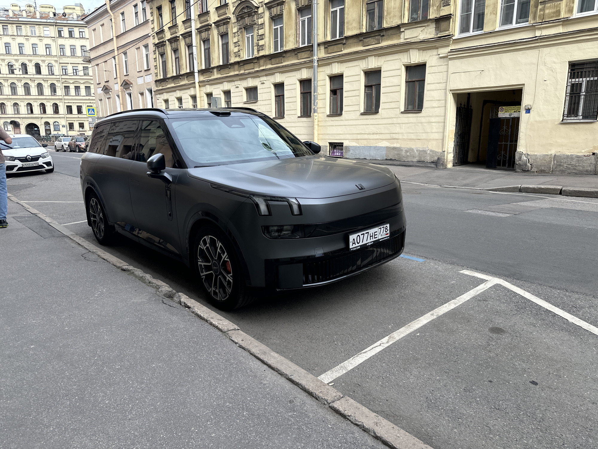 A matte gray Lynk & Co. 09 parked on a city street in Saint Petersburg, Russia. The car is positioned in a parking space marked by white lines on the asphalt. The license plate reads "A077HE 778 RUS". Buildings with varying architectural styles form the backdrop, with light-colored facades and windows. A pedestrian is partially visible on the left side of the frame. The weather appears overcast. An entrance to a building is visible behind the car on the right.
