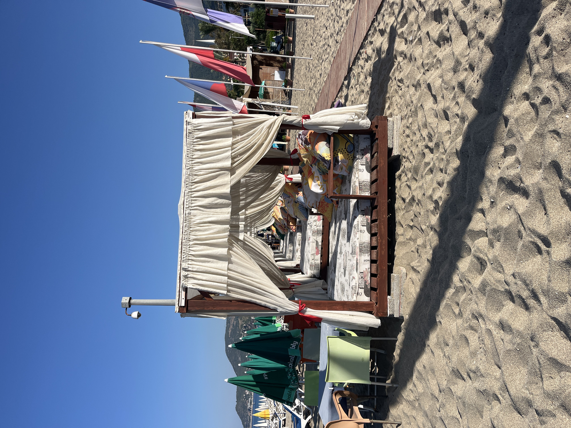 The image displays a sunny beach scene in Alanya, Turkey, featuring a row of wooden cabanas with off-white fabric drapes. The original image is rotated, presenting the scene sideways.
In the foreground, a series of open-sided cabanas are visible, each constructed with dark wooden frames and adorned with gathered white fabric drapes tied back with red ribbons. Inside the cabanas, plush mattresses and cushions with patterned designs, including sketches of buildings and partially visible text like "Club," are arranged for relaxation. A few indistinct figures can be seen lounging within some of the cabanas.
To the left of the cabanas, several green beach umbrellas, some partially folded, are set up alongside light green plastic chairs and other beach furniture. Text on one green umbrella partially reads "Probably" and "#P...bably". Further to the left, the calm blue sea is visible in the distance.
The ground is covered in light-colored sand, showing numerous footprints and casting long shadows from the cabanas, indicating either late morning or early afternoon. A narrow wooden boardwalk runs along the sand to the right of the cabanas.
In the background, tall flags with red, white, and possibly blue colors, reminiscent of national flags, are flying from poles. Behind these flags, lush green foliage and distant mountains rise under a clear, bright blue sky. A security camera is mounted on a metal pole attached to the frame of the leftmost cabana. The overall atmosphere suggests a serene and leisurely beach environment.