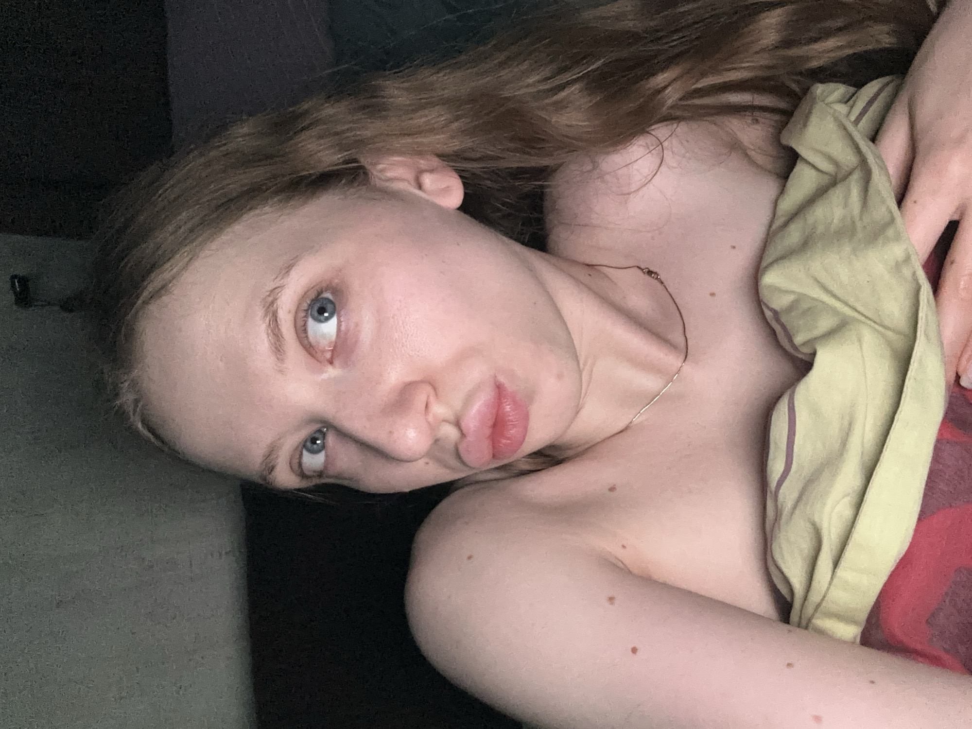 A young woman is depicted in a close-up, horizontal orientation, lying down indoors, likely in a bedroom in Chaikovsky, Russia. She has long, wavy reddish-brown hair that cascades around her head. Her blue eyes are open and directed towards the viewer, and her lips are slightly puckered in a pout. Her fair skin shows a few scattered moles or freckles on her neck and shoulder. She is wearing a thin, delicate chain necklace.
A light green or yellowish fabric, possibly a blanket or sheet, covers part of her body, revealing her bare shoulders and chest. Beneath this fabric, a darker reddish material is visible. The background is dark and indistinct, suggesting a dimly lit room or low light conditions, possibly evening or night. No specific objects are clearly discernible in the background. The overall mood conveyed by her expression is somewhat playful or posed. There is no visible text in the image.