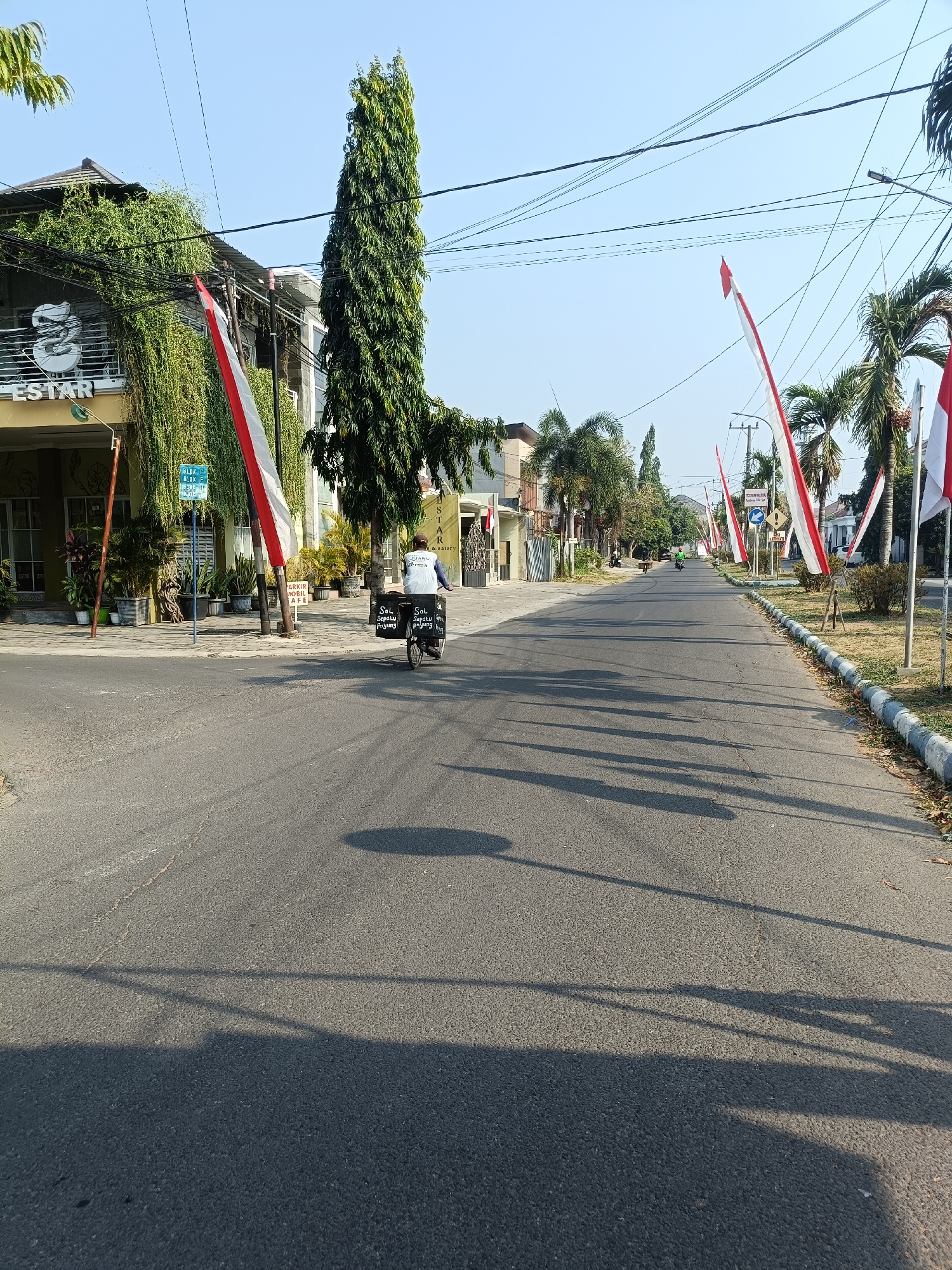 A street scene in Kediri City, Indonesia, features Indonesian flags lining the road. A man on a bicycle with two black boxes on the back is moving away from the camera. The boxes have white lettering. The road is paved and has tire marks and cracks. Shadows of trees and poles are visible on the road. Buildings with greenery are on the left side of the street, and palm trees are on the right. The sky is clear and blue.