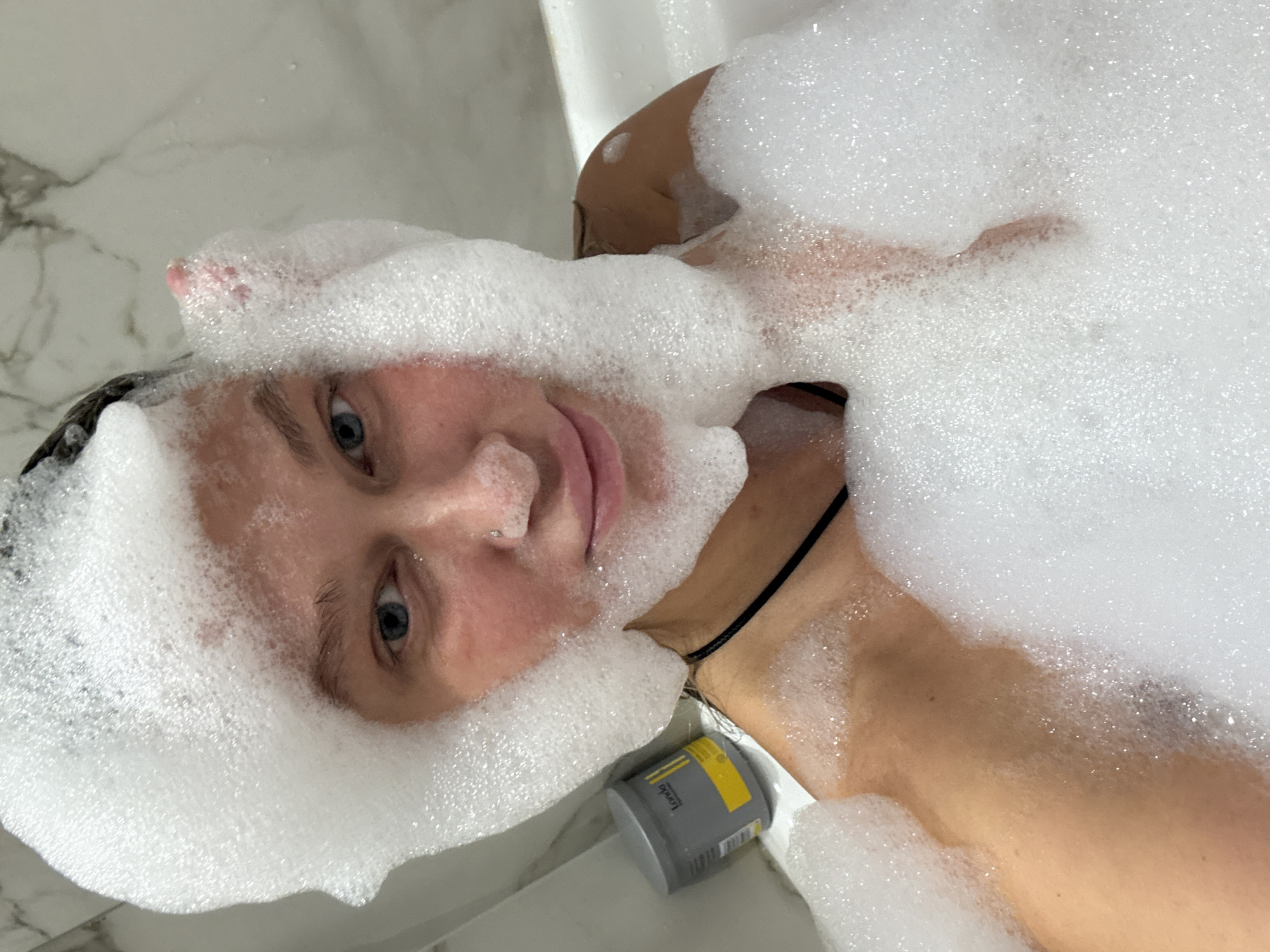 A close-up, rotated portrait shows an individual submerged in a bubble bath within a white bathtub. The person, appearing to be a young adult with light skin and blue eyes, looks directly at the camera. Their face, partially covered by abundant white foam, is positioned to the left side of the frame, with the rest of their body extending towards the right, also largely obscured by bubbles. The skin visible above the foam appears wet.
The setting is a modern bathroom, indicated by white, possibly marble-patterned, tiled walls visible in the background. A black strap, possibly from swimwear or a necklace, is visible around the person's neck. In the bottom right corner, a grey and yellow container of a bath product is partially visible, resting on the edge of the tub. The brand name "Lovob" (or similar, with "Lov" clearly seen) is printed on the label, along with other small, illegible text and what appears to be a barcode.
The image captures a moment of relaxation during a bubble bath, with soft, even lighting suggesting an indoor environment. The person's expression is neutral, engaging directly with the viewer. The context provided indicates the location is Perm, Russia.