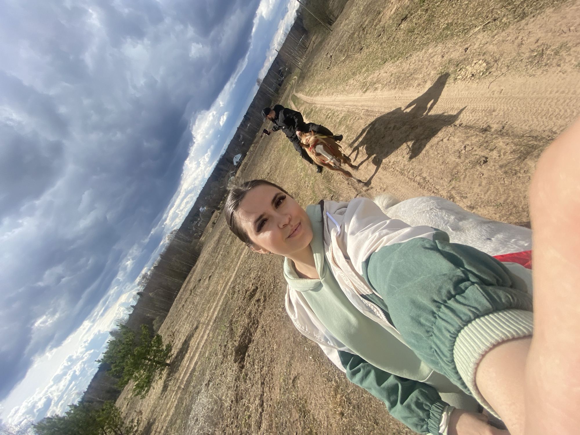 The image is a selfie taken by a woman on horseback. In the foreground, the woman is wearing a green and beige jacket. Behind her, another person is riding a horse on a dirt path. The path leads into the distance, where trees and buildings can be seen. The sky is cloudy. The location is Zhigalki, Russia.