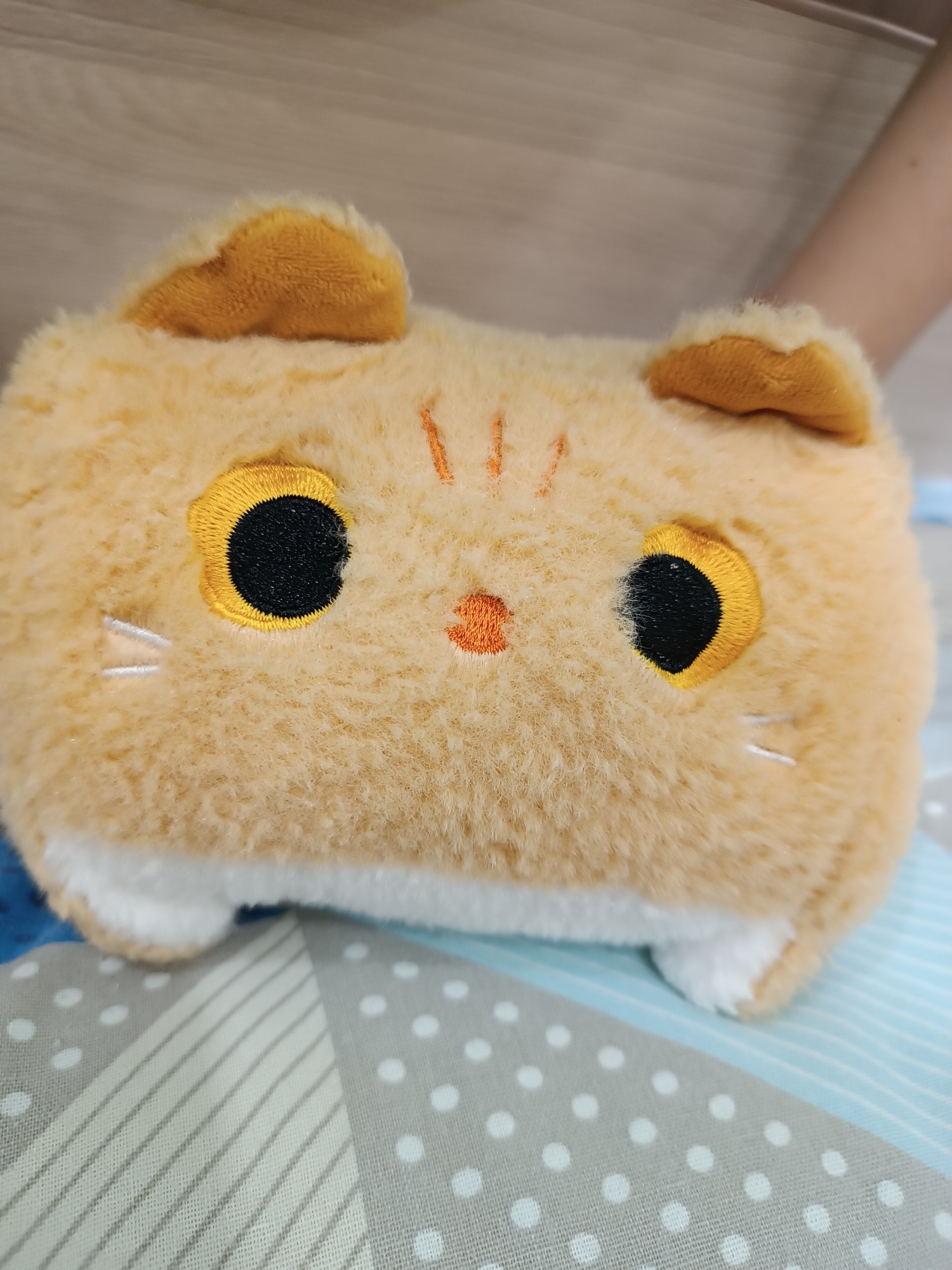 A close-up shows a plush, rectangular orange cat toy with embroidered features. It has two black, round eyes with yellow outlines, a small orange nose, three orange stripes on its forehead, and white whiskers. Two orange ears are attached at the top. The toy's lower body and paws are white. It rests on a bedspread with a geometric pattern of gray triangles, white dots, and light blue stripes. A partially visible arm is in the background, on the right. There is no evidence the image was taken in the city of Chaikovsky, Russia.