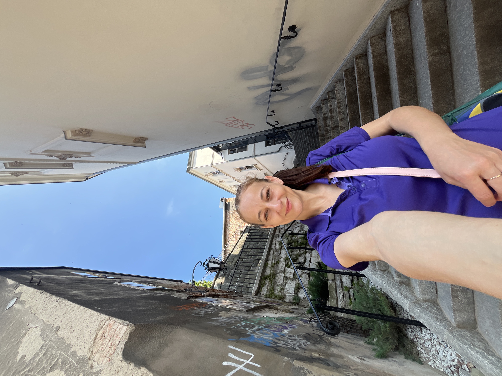 A woman takes a selfie on a narrow, steep stone staircase that runs between two buildings. The building on the left is dark gray with graffiti. The building on the right is a light, creamy color and also has graffiti. The sky is clear and blue. The woman is wearing a purple polo shirt and a light pink strap of a bag is visible across her shoulder. She is smiling and looking at the camera. The staircase has a black metal railing on both sides and there are stone steps with some gravel and small plants visible on the side. A decorative lamp is mounted on the left building.