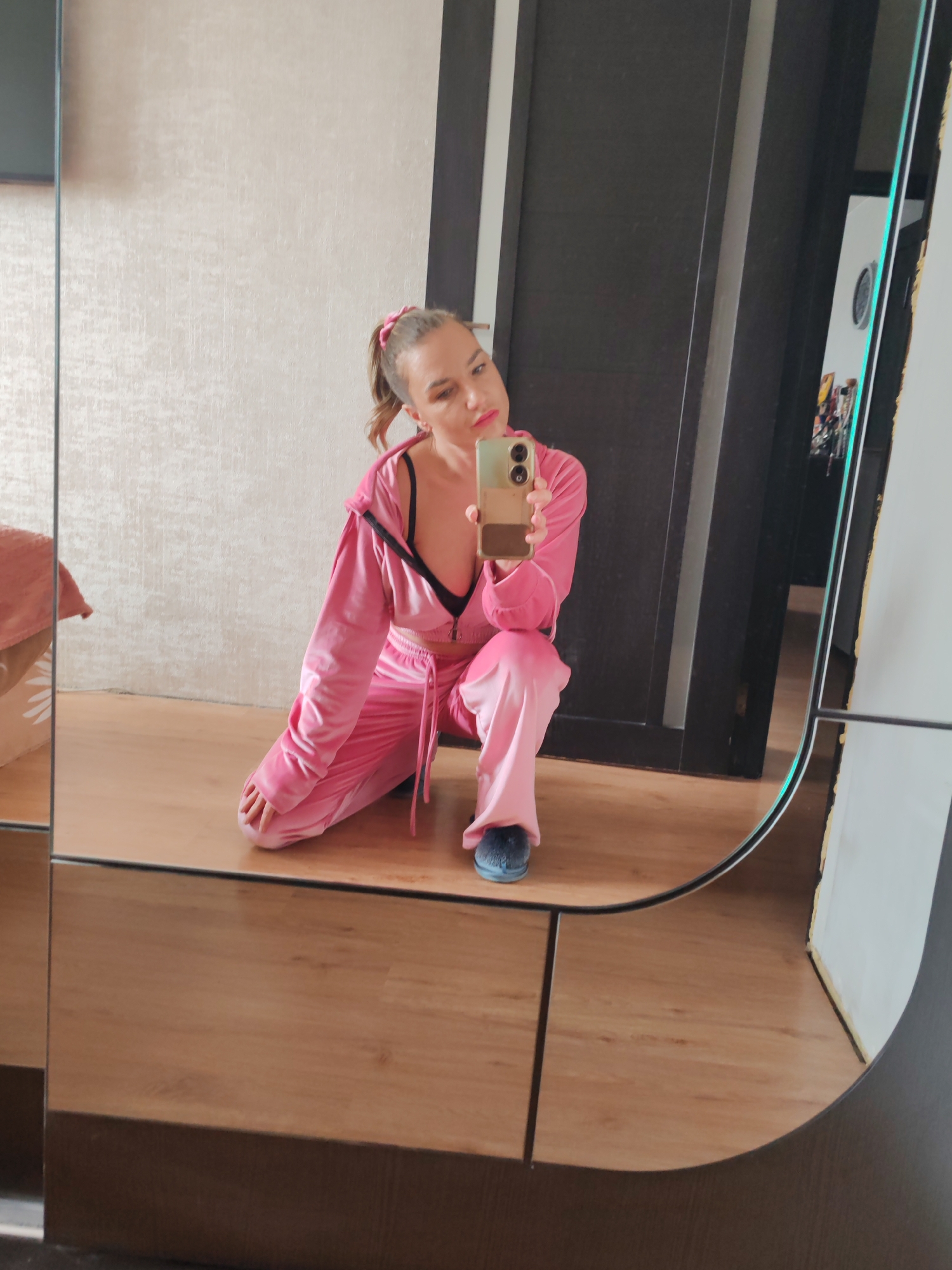 A woman in a pink velour tracksuit kneels on a light brown, wood-look floor, taking a selfie with her smartphone. The background includes a textured light beige wall, a dark brown door with a vertical light accent, and a white wall reflected in a mirror. There is no visual information to ascertain if this image was taken in the city of Petropavlovsk-Kamchatsky, Russia.