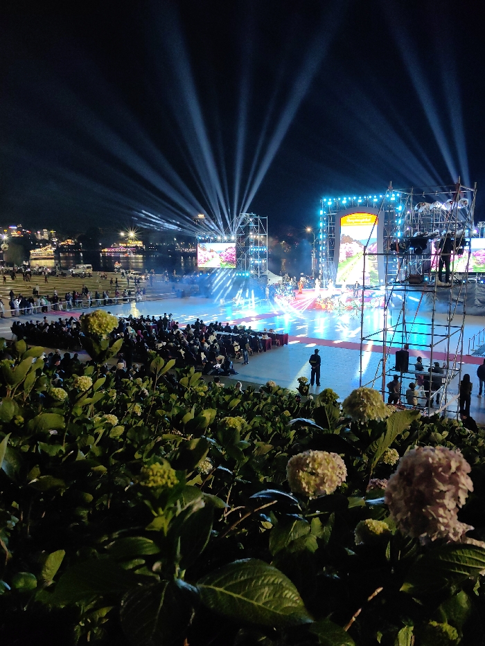 An outdoor nighttime event is captured at Xuan Huong Lake in Da Lat, Vietnam. The foreground is dominated by lush green foliage, specifically hydrangea bushes with some light-colored blossoms and buds. Beyond the foliage, a large, brightly illuminated stage structure is positioned near the lake. This stage features multiple large LED screens displaying scenic imagery, flanked by vertical truss structures supporting additional lighting and screens. Performers, dressed in what appear to be traditional costumes, are visible on the stage floor, which is bathed in blue and white light patterns. Numerous bright white searchlight beams project upwards and outwards into the dark night sky from behind and around the stage.
An audience is seated in rows of chairs facing the stage, occupying a paved area. Further back on the left, additional individuals are standing or walking near the lake's edge. On the right side of the stage, a tall scaffolding structure is present, with at least one individual standing on an upper platform, possibly operating equipment. A man in a dark uniform stands upright in the midground, facing the stage. In the background, the expanse of Xuan Huong Lake is visible, reflecting some of the ambient light. The opposite bank of the lake is lined with numerous city lights, including streetlights, illuminated buildings, and lights from boats on the water, indicating an urban setting. The overall scene is characterized by vibrant artificial illumination against the dark night.