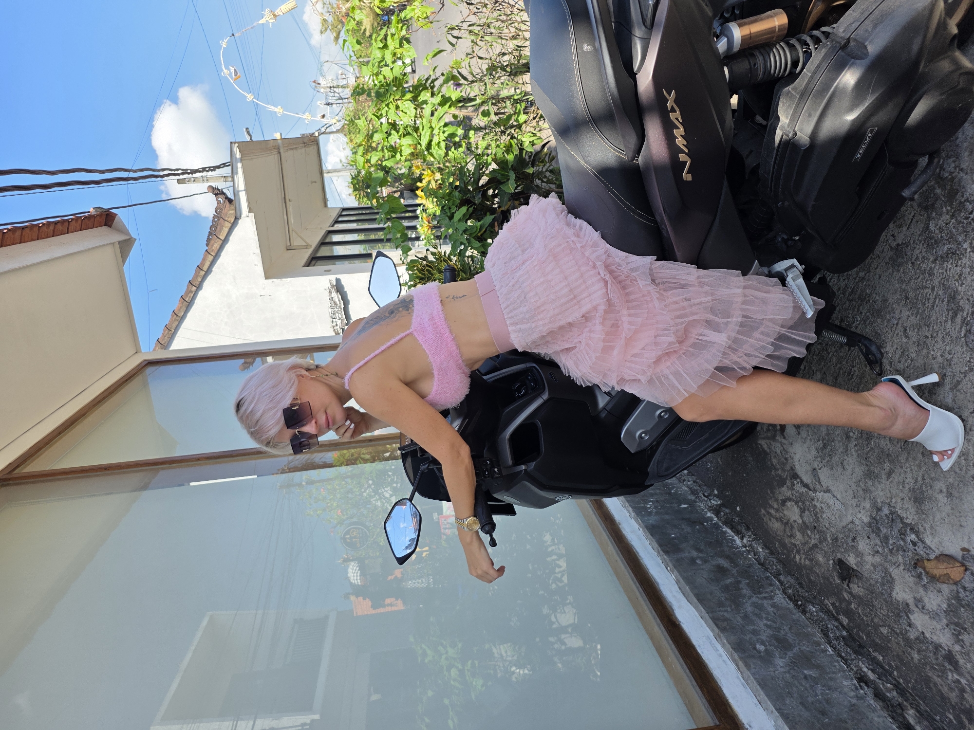 A woman with short, pink hair and sunglasses sits on a black scooter, identified by the "NMAX" logo on its side. She is wearing a pink, fluffy bralette and a layered, pink tulle skirt. Her left hand rests on the scooter's handlebars, and her right hand is near her face. She is wearing white heeled mules. The scooter is parked on a grey, textured surface in front of a building with a large window. Behind the building, lush green foliage and trees are visible under a clear blue sky with a few white clouds. An arched decorative structure with what appears to be straw is also visible in the background. The scene is outdoors on a sunny day, suggesting a location in the city of North Kuta, Indonesia.