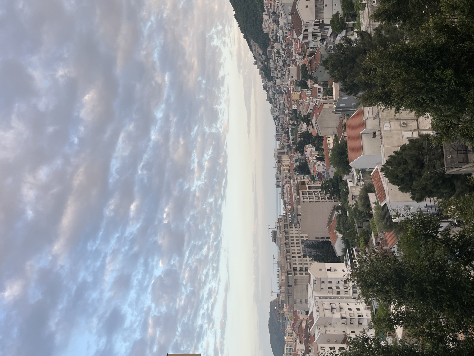 The image depicts a cityscape in Budva, Montenegro. A mixture of residential and commercial buildings extends along the coast and up a hillside, characterized by white facades and red-tiled roofs. The sky is filled with textured clouds. The sea is visible between the buildings. Trees are present in the foreground, obscuring some parts of the buildings. Construction cranes are visible in the distance. No people are visible in the frame.