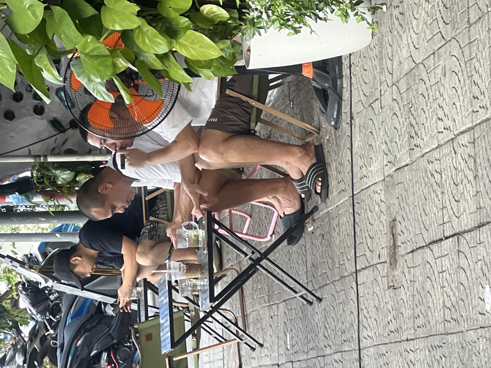 Three men are seated at small, black, folding tables outside a building, likely a cafe or restaurant, on a paved outdoor area. The man on the left, wearing a black t-shirt and black baseball cap, is looking at his phone. The man in the center, wearing a white t-shirt, is also looking down, possibly at his phone. The man on the right, wearing a white t-shirt and dark shorts, is holding a cigarette to his mouth. In front of the men are drinks, including a glass of iced tea. A black oscillating fan with orange blades is positioned behind the man on the right, partially obscured by lush green foliage. In the background, several motorcycles are parked. The pavement is made of grey square tiles with a decorative pattern. The scene is consistent with an outdoor seating area in Đà Nẵng, Vietnam, likely during the daytime due to the ambient light.