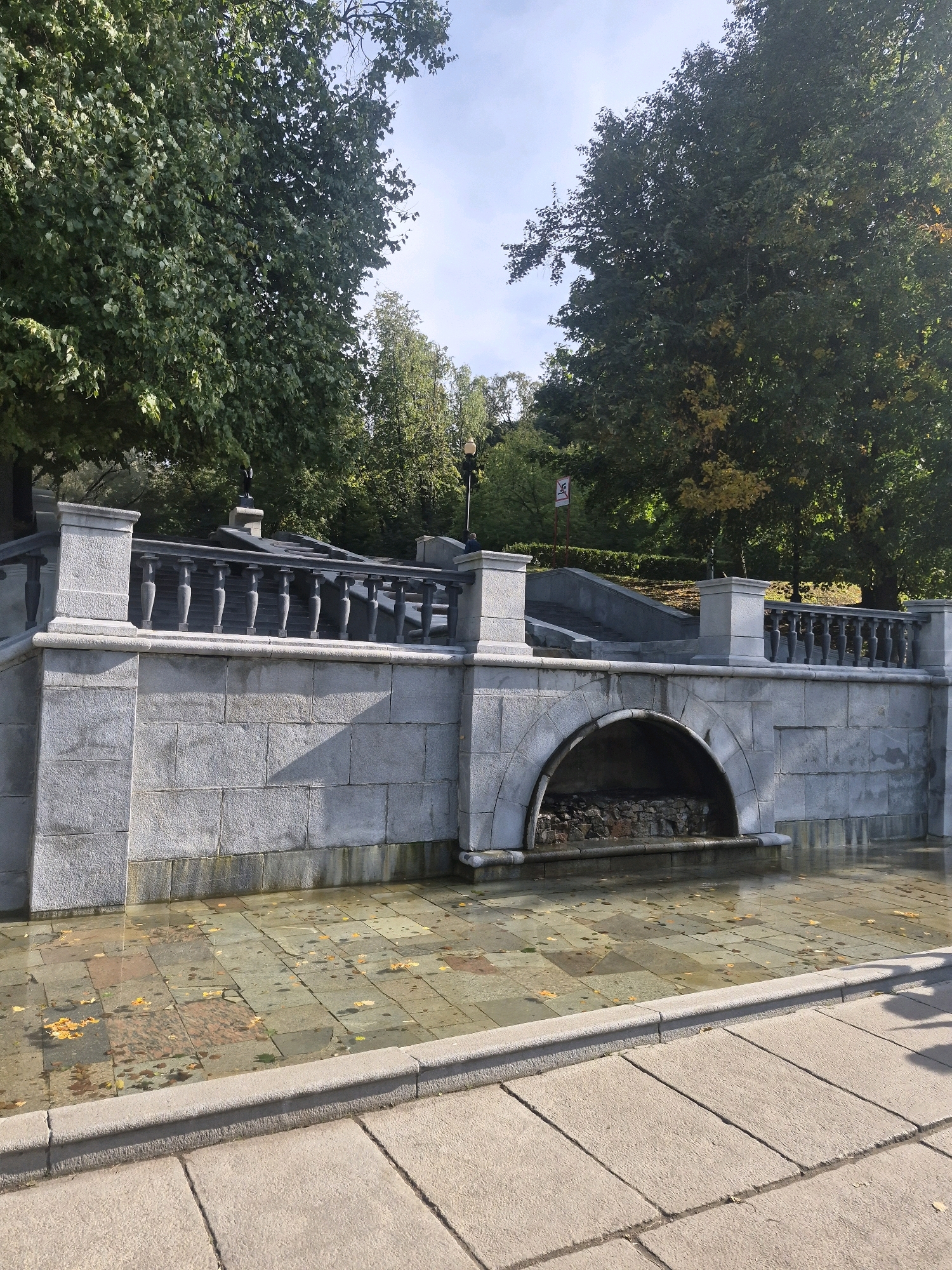 The image shows a paved area in front of a stone structure with a balustrade and an arched niche. Water covers the paved area in front of the structure, and fallen leaves are scattered on the surface. Trees with green and yellow foliage are visible in the background. The location is the Alexander Garden in Moscow, Russia, specifically near the fountains. The scene appears to be a sunny day with a clear sky. No people are clearly visible.
