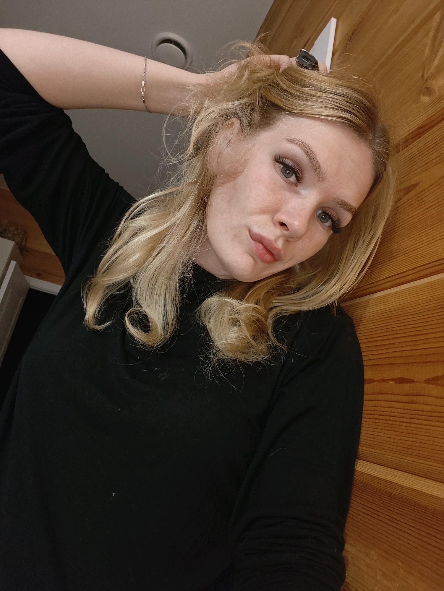 A young woman with blonde hair is shown from the chest up, leaning against a wooden wall. She is wearing a black, long-sleeved shirt. Her left arm is raised, and her hand is in her hair. She is wearing a delicate silver bracelet on her left wrist. Her head is tilted, and she is looking towards the camera with a neutral expression. The background consists of a warm-toned wooden wall. The lighting is soft, and the image appears to be taken indoors. There are no discernible city landmarks or specific geographical features of Русский Вожой, Russia, visible in the image.