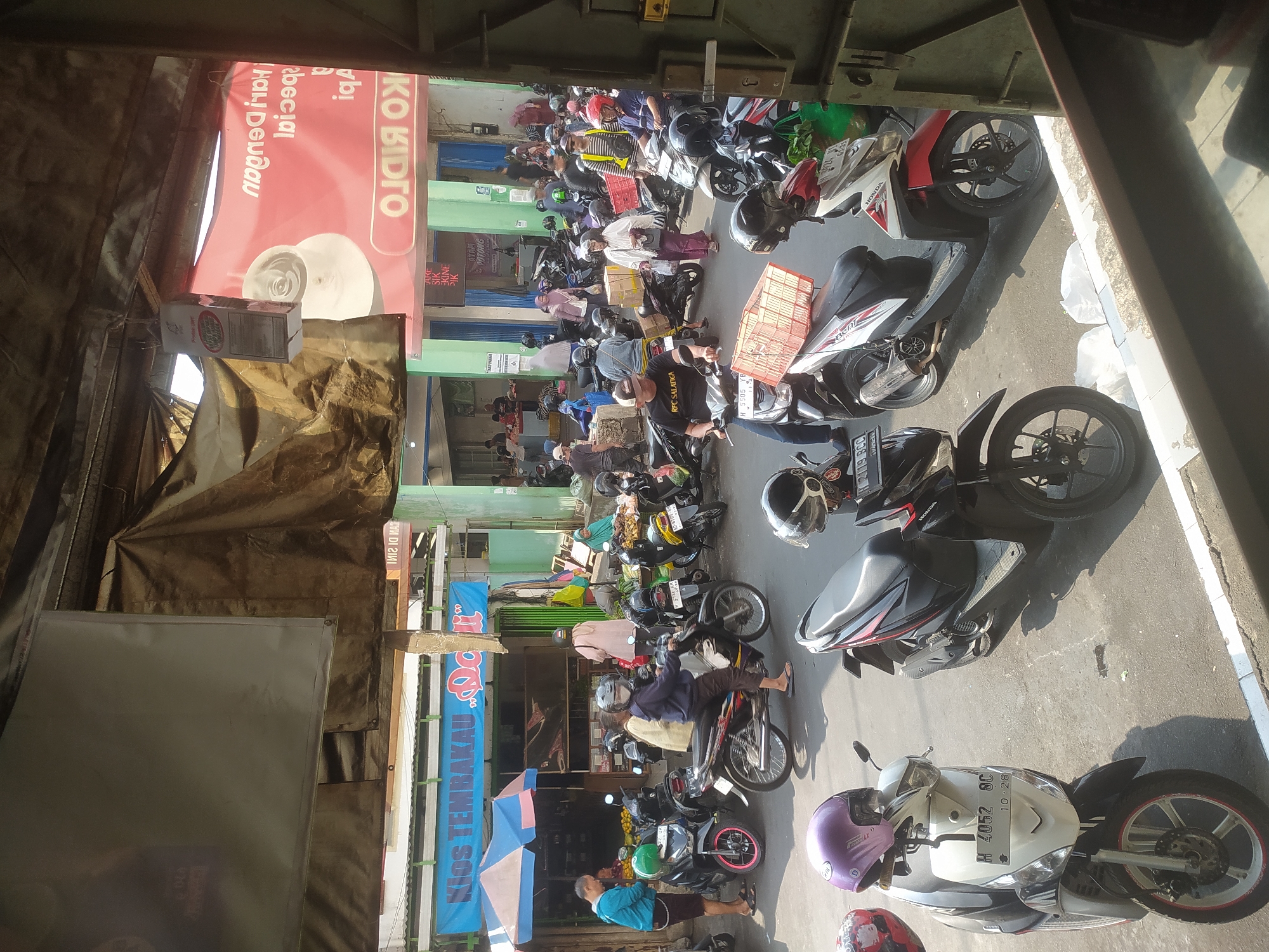 The image depicts a busy outdoor market scene in Salatiga, Indonesia, likely a street market. The foreground features several parked motorcycles and scooters, some with helmets placed on them. In the midground, people are walking and interacting, with more motorcycles and stalls visible. One stall prominently displays a blue banner with "Kios Tembakau Dolii" written on it, indicating a tobacco shop. Further in the background, there are more market stalls, buildings with green and red facades, and a crowd of people. Some individuals are seen carrying goods, and several motorcycles are in motion. The overall atmosphere suggests a lively and active commercial area.