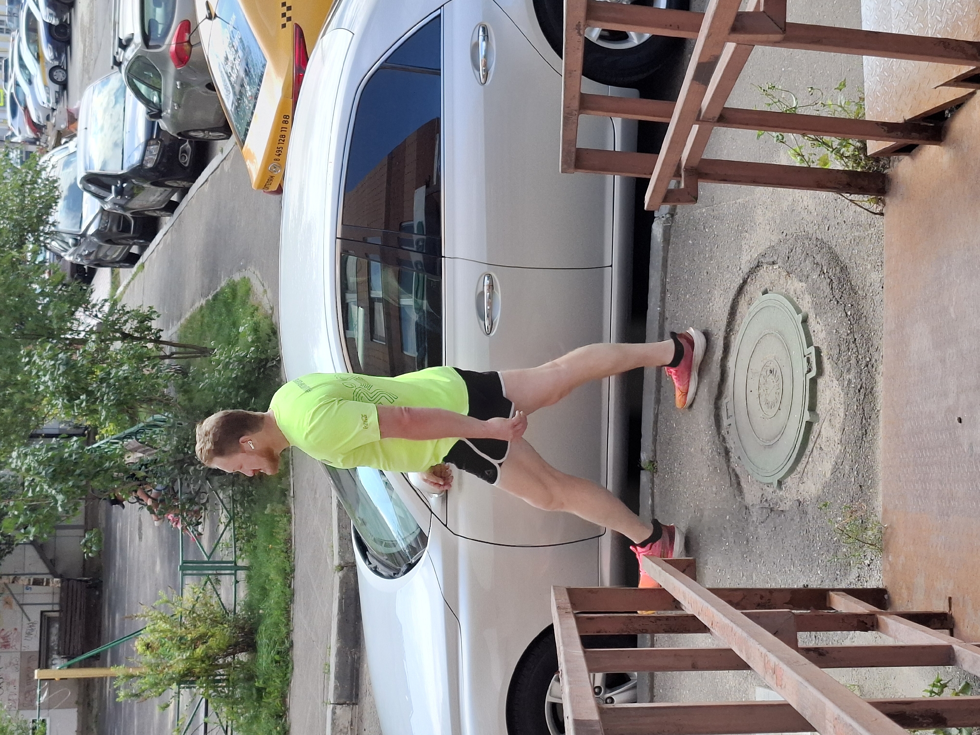 A man in a neon green t-shirt and black shorts with white trim is stretching his legs on an outdoor concrete walkway in front of a silver sedan. He is wearing white earbuds and pink running shoes. The man is facing towards the right side of the frame. Behind him, there is a silver sedan with tinted windows. The walkway is cracked in places and there is a circular manhole cover in the foreground. To the left of the man, there is a rusted metal railing and steps leading down. In the background, there are parked cars and trees. The scene appears to be in a residential urban area.