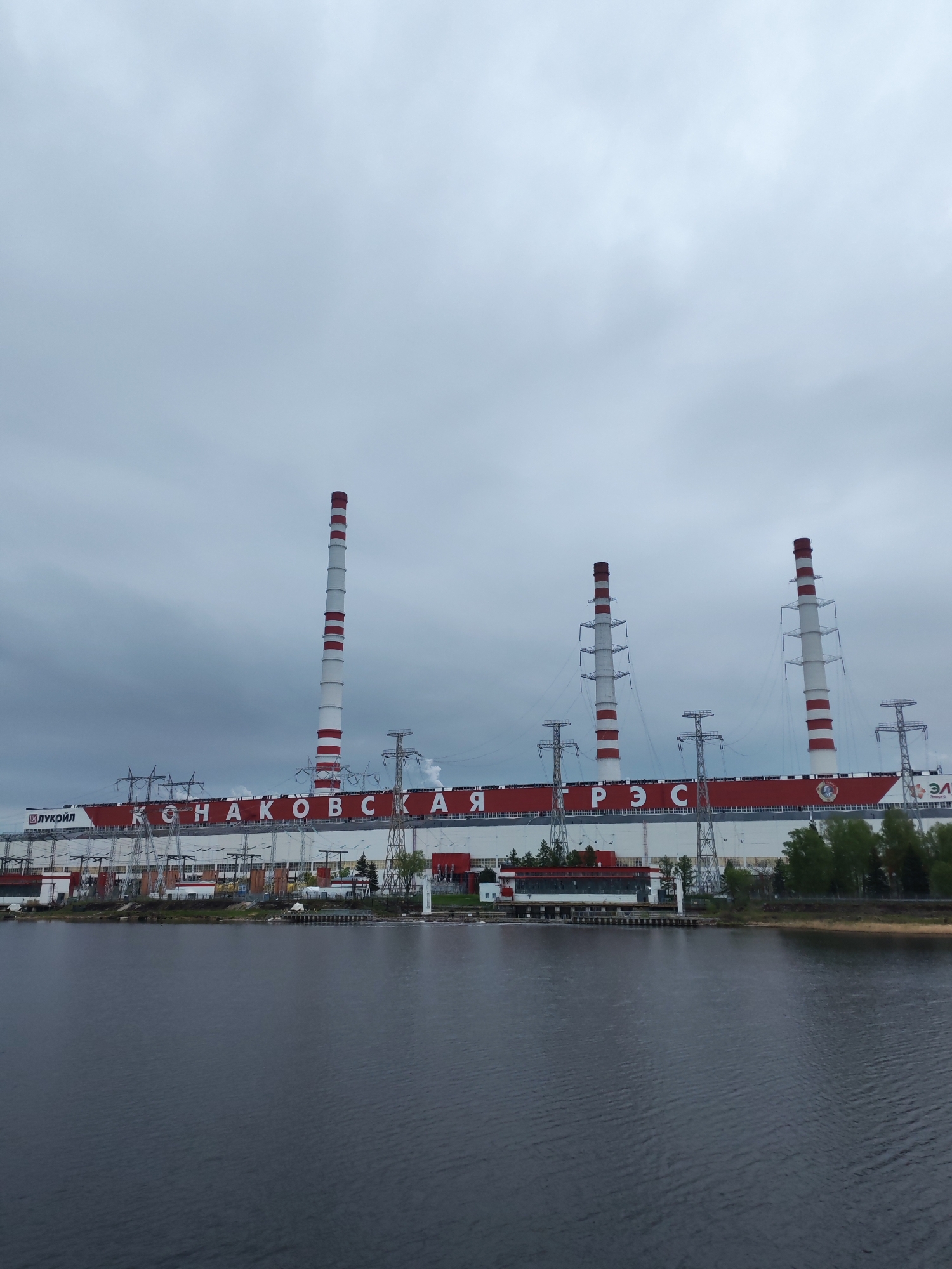 The image depicts the Konakovo Power Station in Konakovo, Russia. The station is a large industrial complex with multiple tall smokestacks striped in red and white. Power lines extend from the station, supported by metal towers. The name of the station, "Конаковская ГРЭС", is written in large Cyrillic letters across the building. A logo for "ЛУКОЙЛ" is also visible. The station is situated on the bank of a body of water, possibly a river or lake. The sky is overcast.