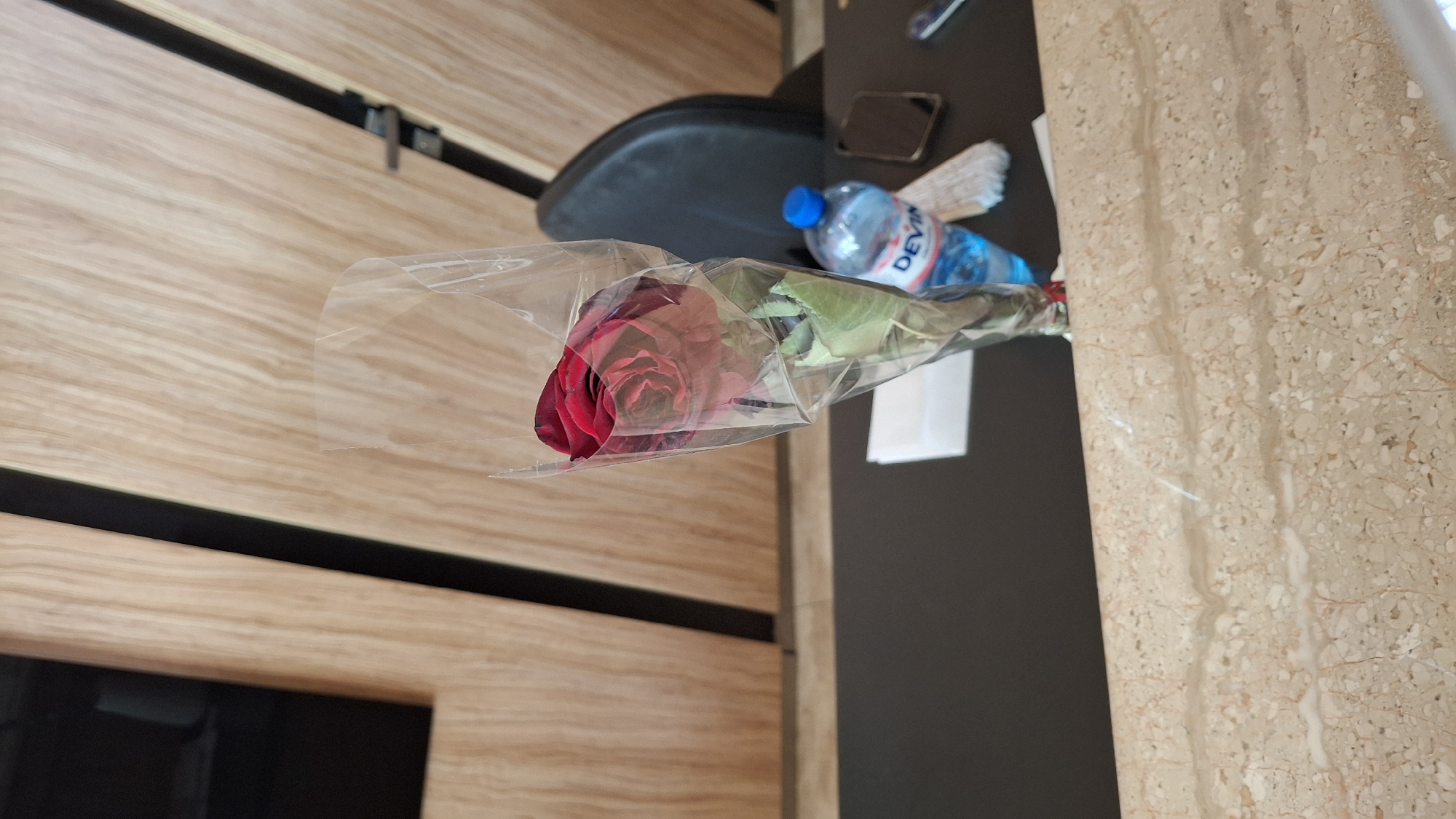 A single red rose, wrapped in clear plastic, is presented in a close-up shot. The rose is the focal point, positioned in the center of the frame with its petals unfurled and a deep red hue. It is presented in what appears to be an indoor setting, likely a desk or table, with a dark, flat surface in the foreground and a textured, wood-paneled wall behind. A partially visible black chair is also present in the background. To the right of the rose, a blue bottle of "DEVIN" mineral water is situated, alongside a smartphone and a folded white paper. The scene is lit by overhead lighting, casting subtle shadows and highlighting the textures of the wood and the rose petals. The location in Nesebar, Bulgaria, is not explicitly indicated by any visible landmarks or signs within the image itself.