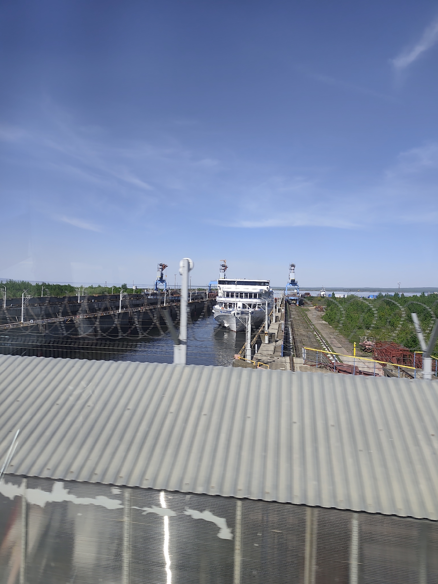 The image shows a river lock with a passenger ship inside, viewed from a slightly elevated perspective, possibly from a building or another ship. The lock is surrounded by industrial structures, including cranes and metal frameworks. The sky is mostly clear with a few scattered clouds. The scene is located in Chaikovsky, Russia, based on the visual characteristics of the environment and structures. A corrugated metal structure is in the foreground and out of focus. Vegetation, including trees and shrubs, is visible on the banks of the lock.
