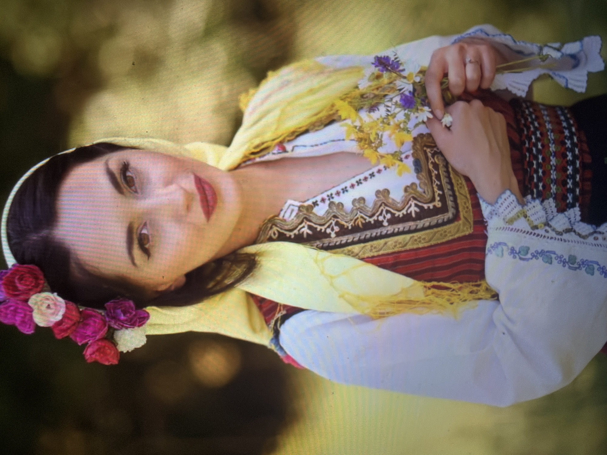 A woman, appearing to be in her 20s or 30s, is centered in a medium shot, looking directly at the viewer with a slight smile. She has dark hair, brown eyes, and is wearing visible makeup, including eyeliner and red lipstick. Her head is adorned with a white band, a yellow scarf draped over her head and left shoulder, and a floral arrangement of pink, red, purple, and white artificial flowers positioned above her left ear. She is dressed in traditional attire consisting of a white long-sleeved blouse with embroidered cuffs and a V-neck featuring cross-stitch details. Over the blouse, she wears a sleeveless garment with vertical red and dark brown stripes, accented with gold and brown embroidery around the neckline and borders. A wide waist sash with geometric patterns in red, white, and dark colors is also visible. Her hands are clasped at waist level, holding a small bouquet of wildflowers, predominantly yellow, purple, and white. A simple ring is visible on her left ring finger. The background is a softly blurred outdoor environment featuring green and yellow foliage, under natural daylight. This scene takes place in the city of Shumen, Bulgaria.