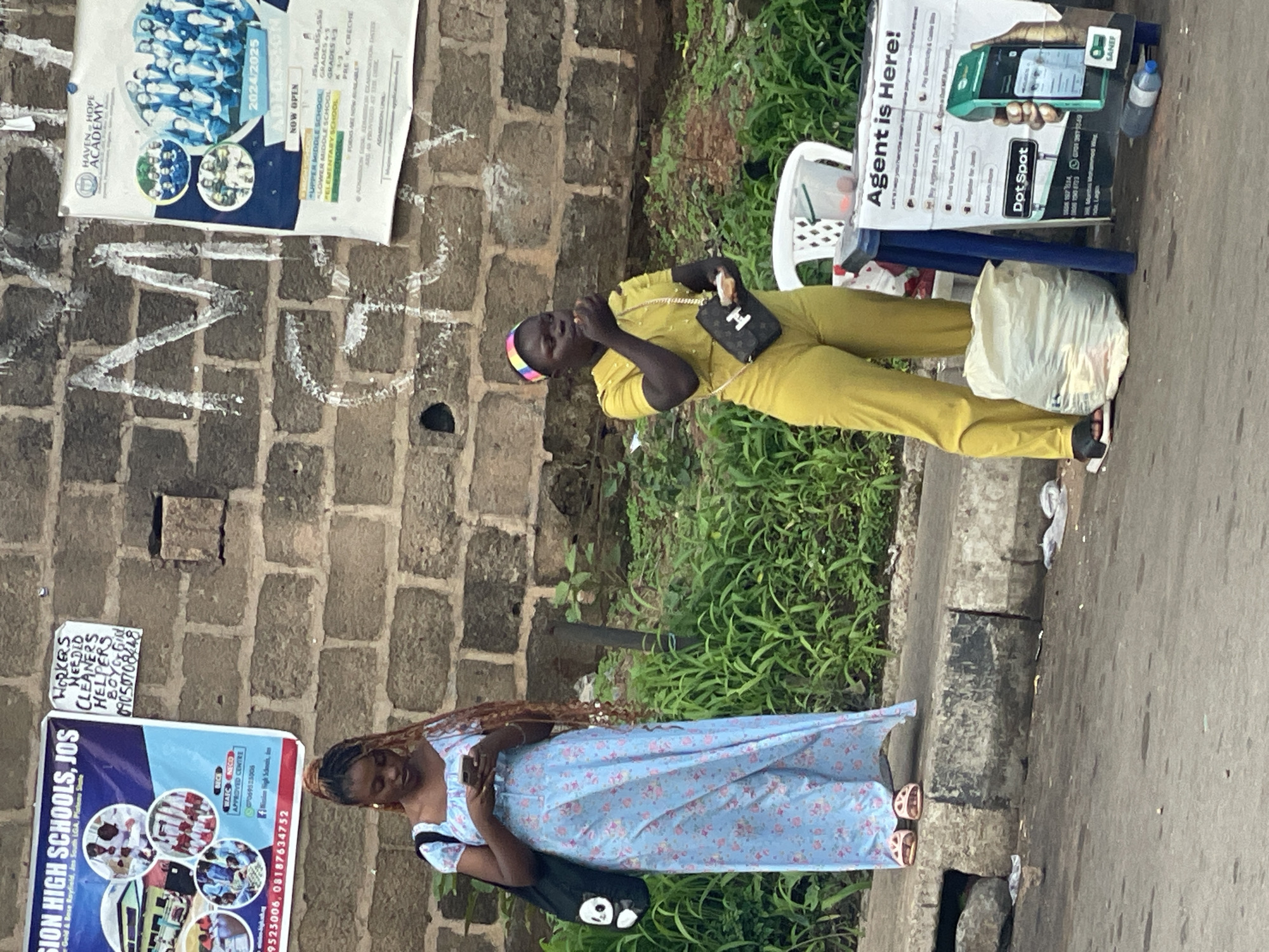 A wide shot captures an outdoor scene in Barakin Lerawi, Nigeria, with a focus on two young women standing near a brick wall plastered with advertisements. The woman on the left wears a long, flowing light blue floral dress and sandals, holding a phone in her hands. A dark tote bag with a panda design hangs from her shoulder. The woman on the right is dressed in a bright yellow jumpsuit and a colorful headband, eating something and carrying a black purse. In the foreground, a white chair and a blue table support a sign for "Dot Spot," an agent service that offers various financial transactions. A large white bag, possibly containing trash, is placed on the ground near the second woman. The environment suggests a street or roadside setting, with a paved area, a curb, and overgrown vegetation visible behind the women. The brick wall exhibits some graffiti.