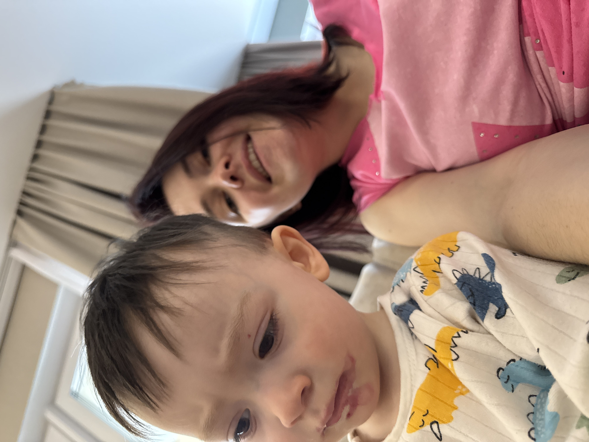 A woman and a young child are pictured indoors. The woman is partially visible with dark hair, a pink shirt, and is smiling. The child is positioned closer to the foreground and appears to have some substance smeared around the mouth. The child is wearing a white shirt with dinosaur patterns. The background includes curtains and what seems to be a window. The photo appears to be taken at an angle. The image does not contain any evidence or indications that would provide the location of Izhevsk, Russia.