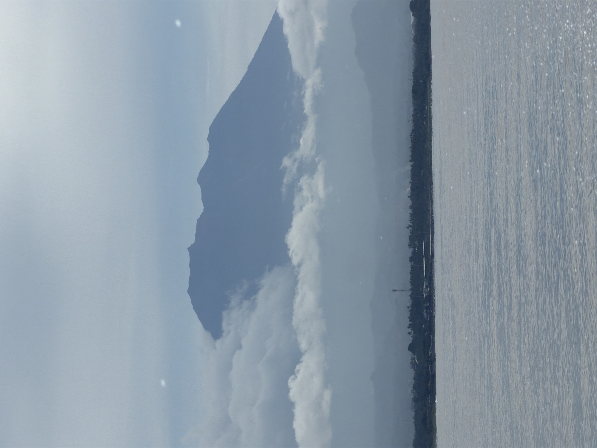 The image shows a body of water with a shoreline along the right side, where some trees and structures are visible. To the left of the shoreline, there's a large mountain partially obscured by clouds. The sky is predominantly cloudy. The location is possibly in Indonesia, but the exact city is unknown.