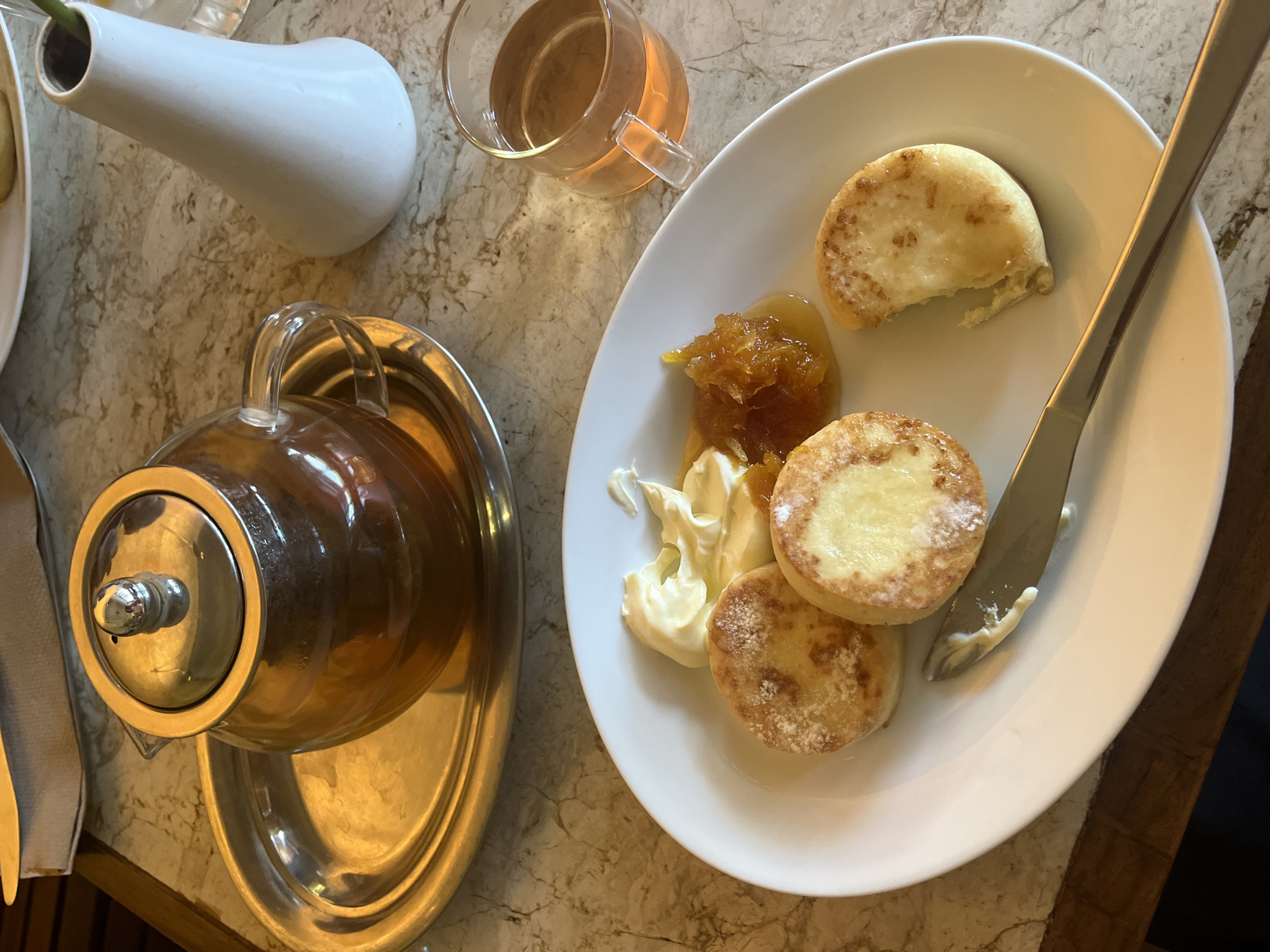 The image shows a tabletop scene of a meal, likely breakfast or brunch. The setting appears to be indoors, on a marble-patterned table. A white, pear-shaped vase sits near a clear glass mug containing a light amber-colored liquid, possibly tea. A silver teapot on a matching silver tray is positioned on the left side. A white plate dominates the right side of the frame, holding three small, round pancakes or syrniki. A bite has been taken from one of them. The syrniki are accompanied by a dollop of white cream and what looks like orange marmalade or jam. A silver knife rests on the plate. The environment suggests a casual or cafe setting. The location in North Kuta, Indonesia is not evident from the image.