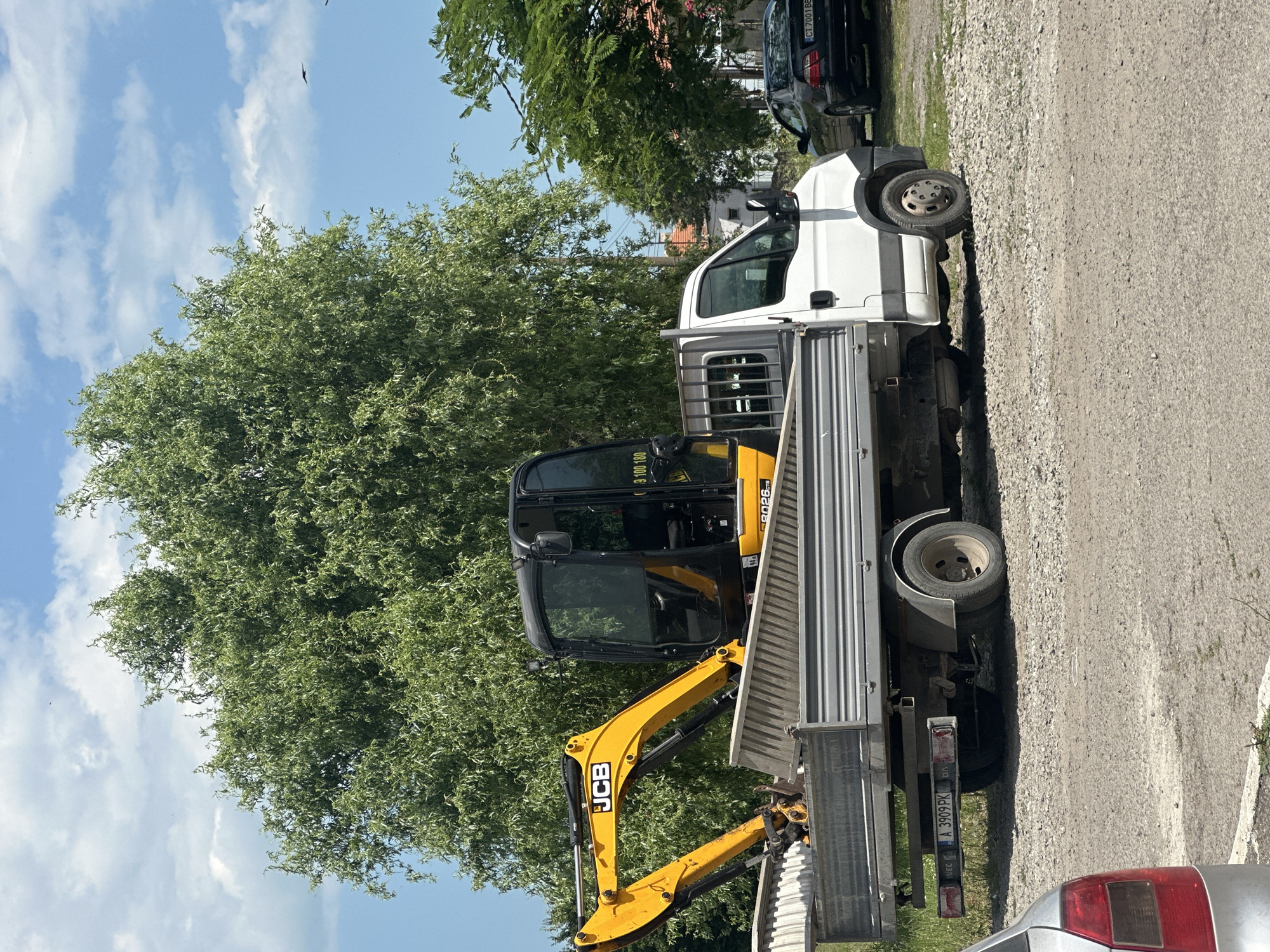 In Burgas, Bulgaria, a yellow and black JCB mini-excavator is loaded onto the flatbed of a white Iveco truck. The truck is parked on a gravel surface, with a large green tree in the background. To the right of the truck, a dark-colored Mercedes-Benz sedan is parked. A portion of a silver car with a red taillight is visible in the lower left corner of the frame. The sky is blue with scattered white clouds. The scene appears to be a sunny day in a somewhat rural or industrial area.